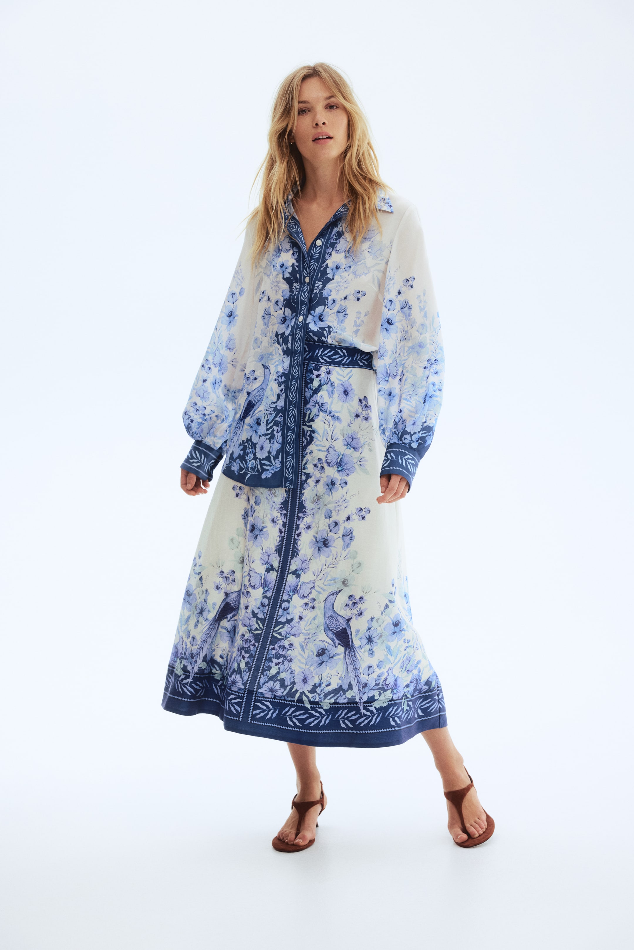 View larger image: A two-piece set, facing forward, features a white base with an all-over blue floral and bird pattern. The button-down shirt is designed with a pointed collar, long, full sleeves ending in wide patterned cuffs, and a matching fabric belt cinches the waist of the flowing midi skirt.