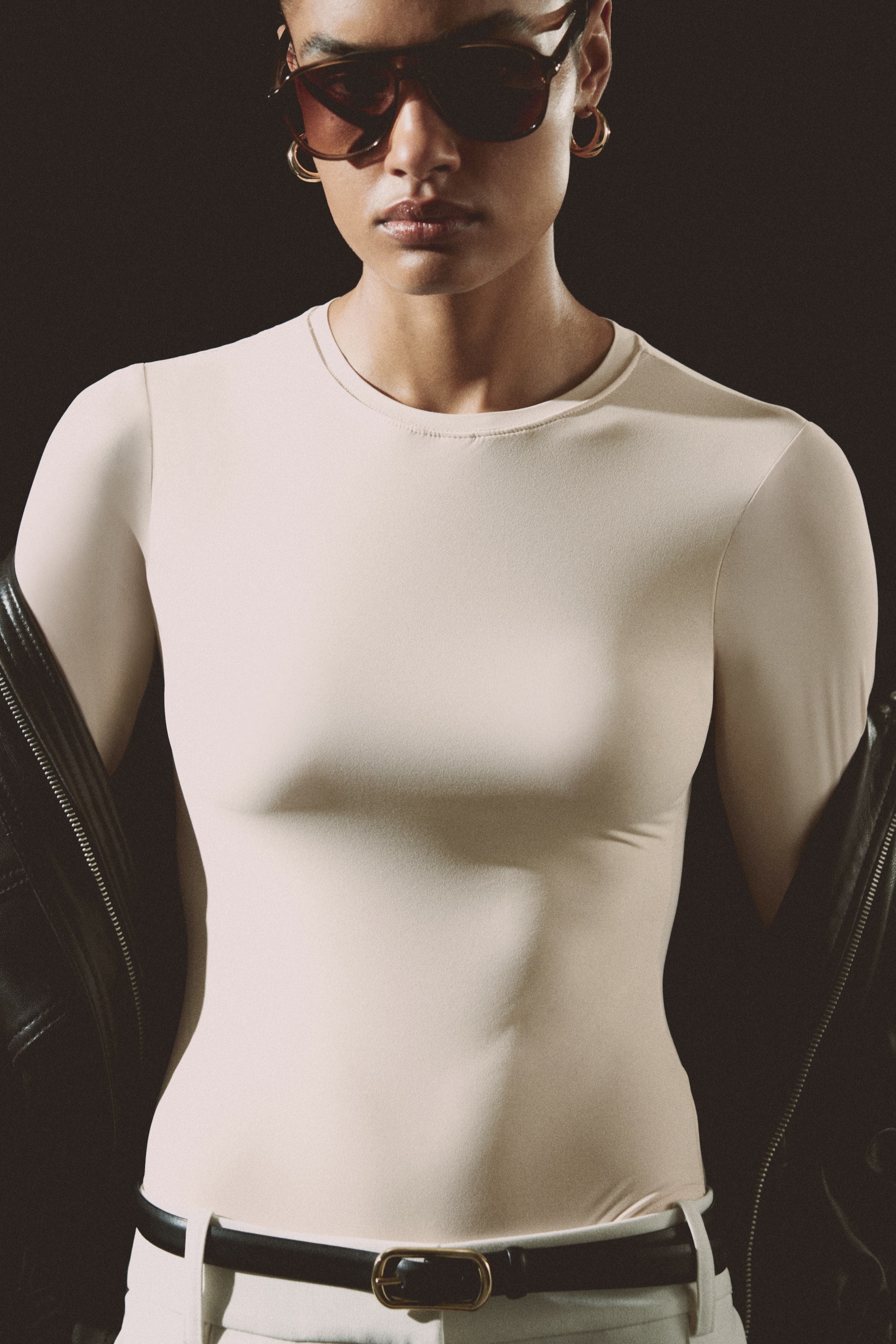 View larger image: A light beige, form-fitting, long-sleeved top featuring a crew neck and a smooth texture. It's shown facing forward, tucked into white trousers with a black belt, and a black leather jacket draped over the shoulders.