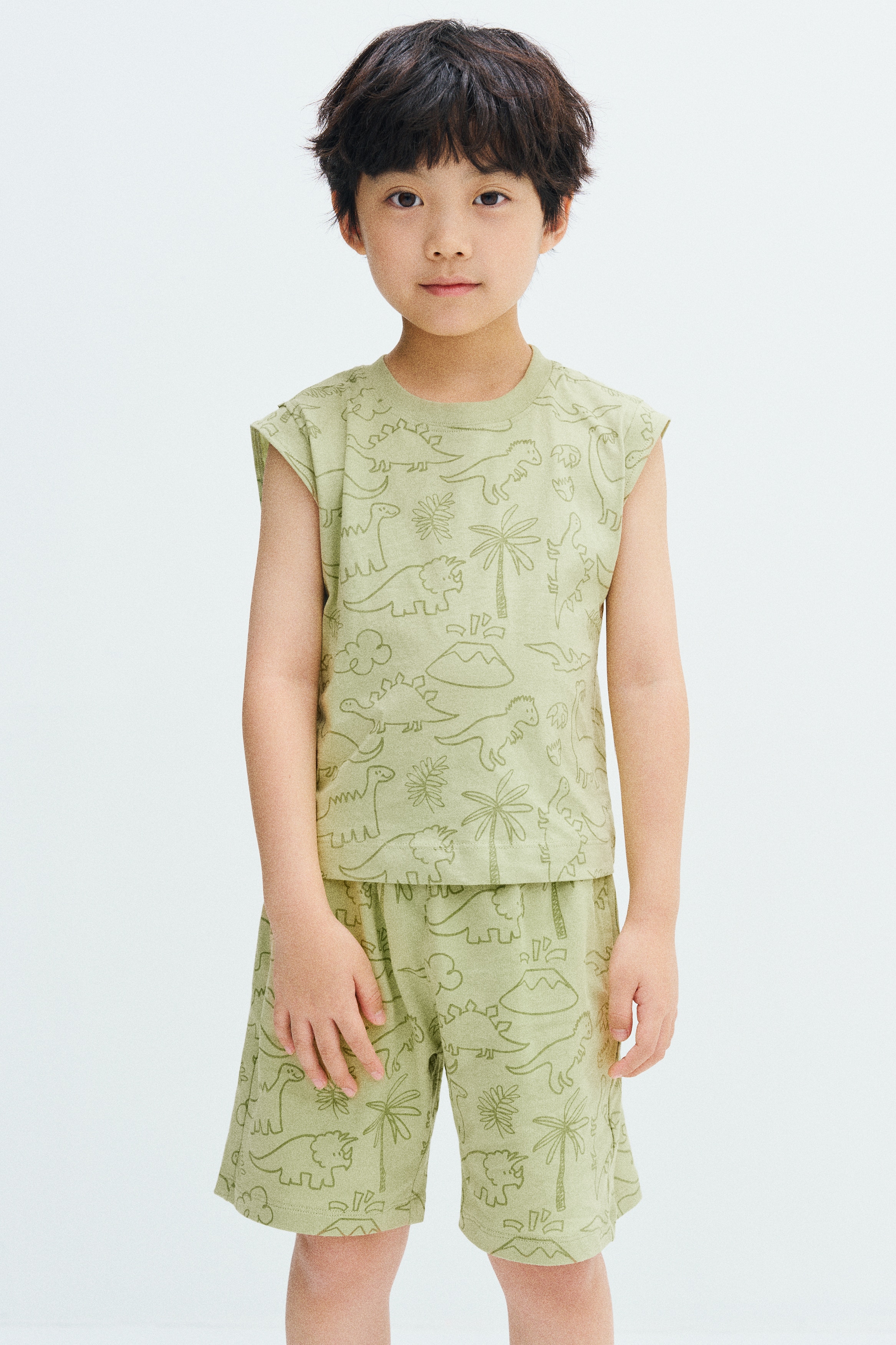 A light green sleeveless top is displayed in front view with an all-over print of olive-green, line-drawn dinosaurs, volcanoes, and palm trees. The top features a ribbed crew neck and matching ribbed armhole trims.
