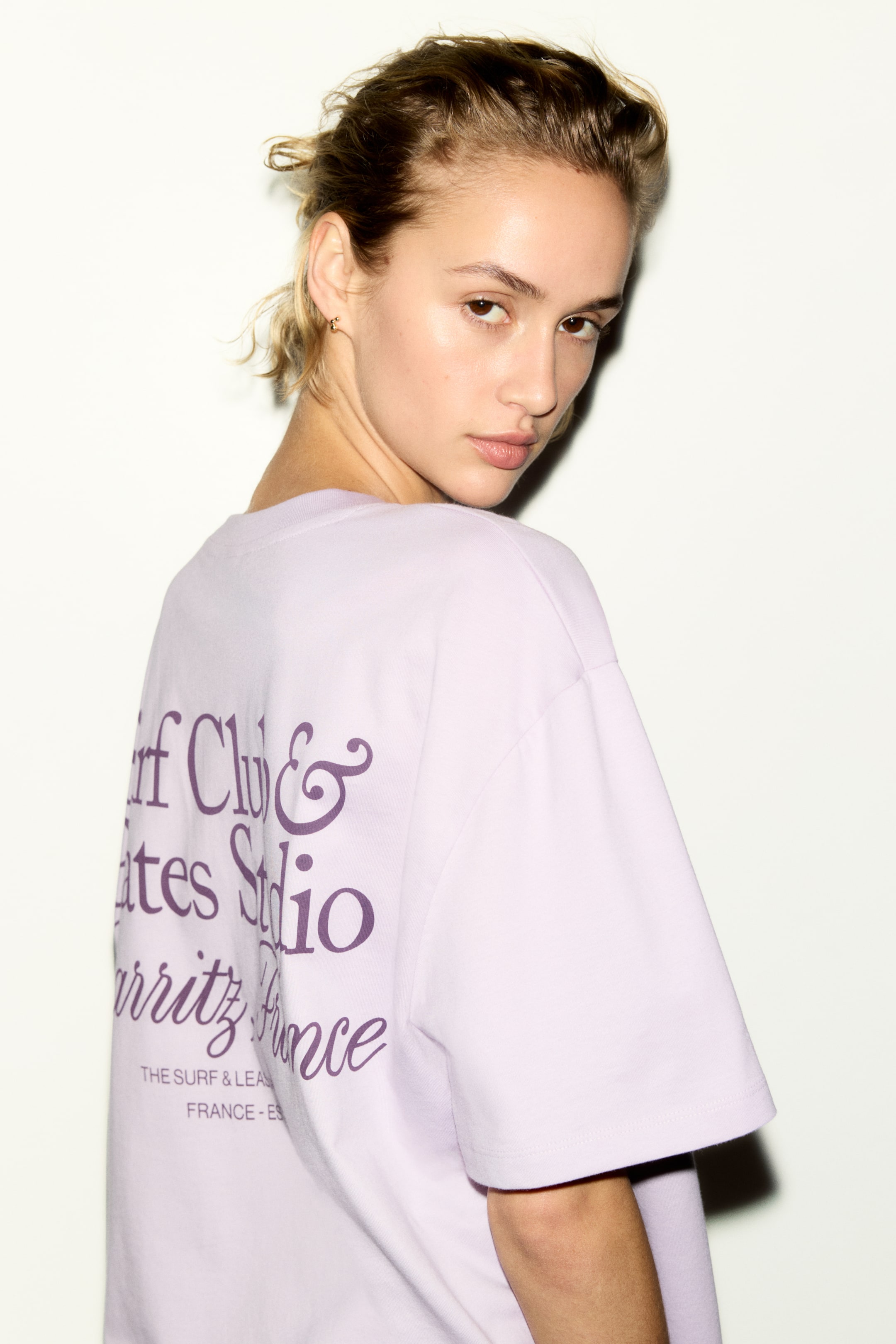 View larger image: A lilac t-shirt is showcased from the back, featuring a large purple graphic print on its reverse. The print displays 'Surf Club & Pilates Studio Biarritz France' in a script font, with 'THE SURF & LEAGUE FRANCE - ES' in smaller text below. The t-shirt is designed with short, wide sleeves.