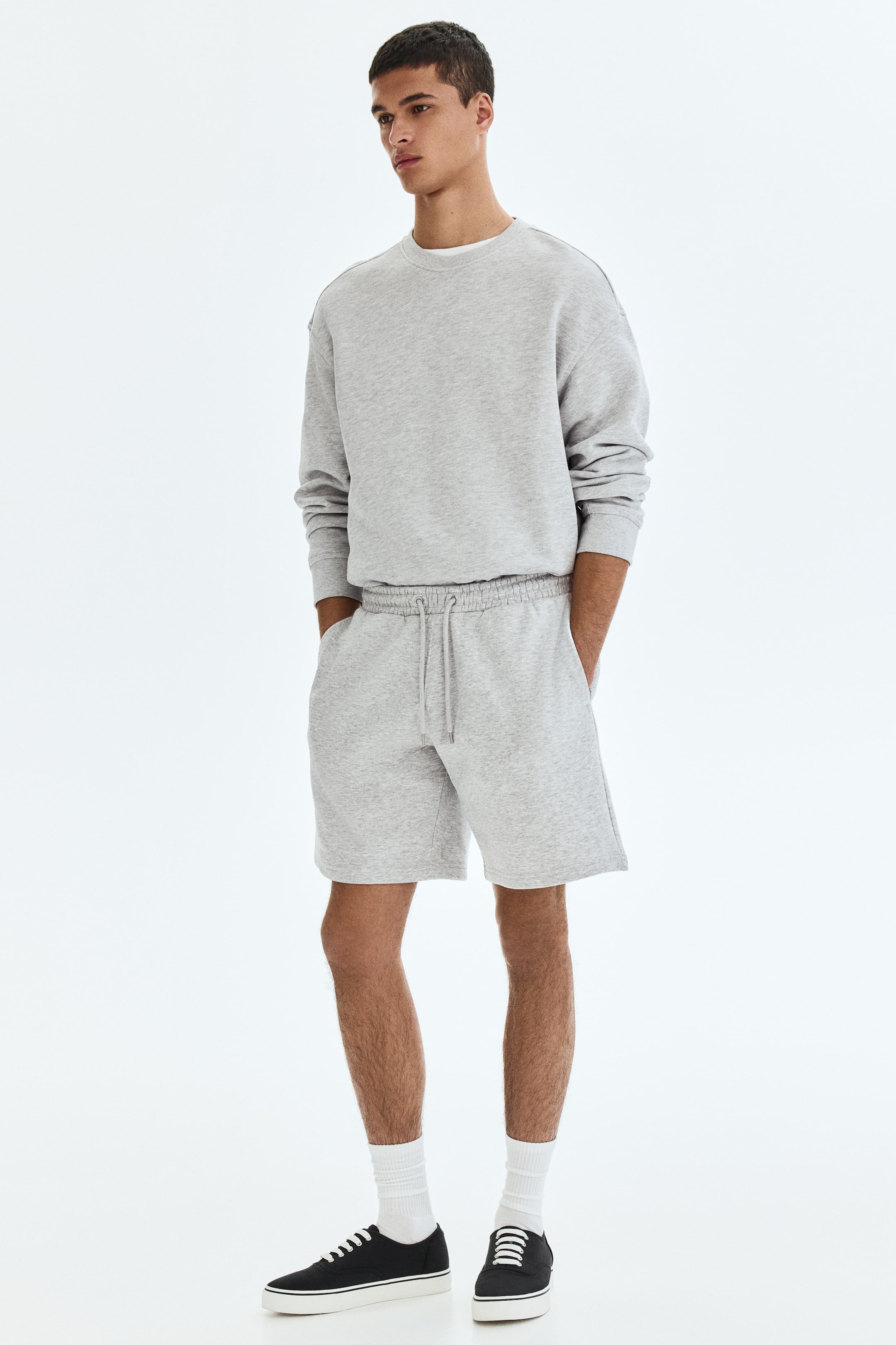 Visa större bild: Ett ljusgrått melerat matchande set, bestående av en oversized crewneck-tröja och shorts, presenteras från en något vinklad frontvy. Tröjan har en ribbad crewneck med en vit t-shirt under, samt nedhasade axlar och ribbade muddar, medan shortsen har en elastisk dragsko i midjan och sidofickor.