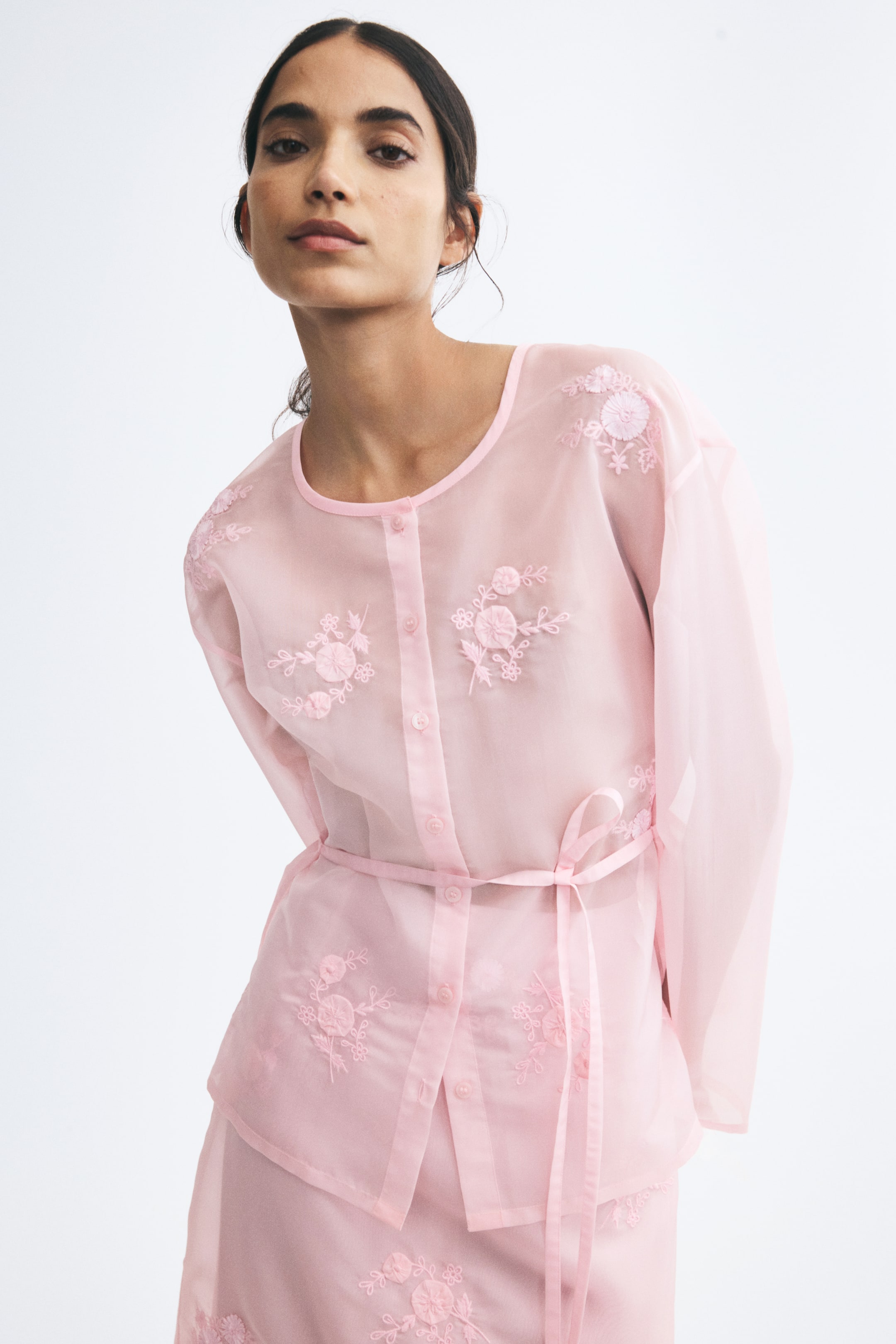 View larger image: An embroidered organza blouse in light pink is being worn by the model, paired with a matching embroidered skirt. The sheer, button-up blouse features delicate floral embroidery on the chest, shoulders, and hem, and is styled with a matching thin tie-belt at the waist.