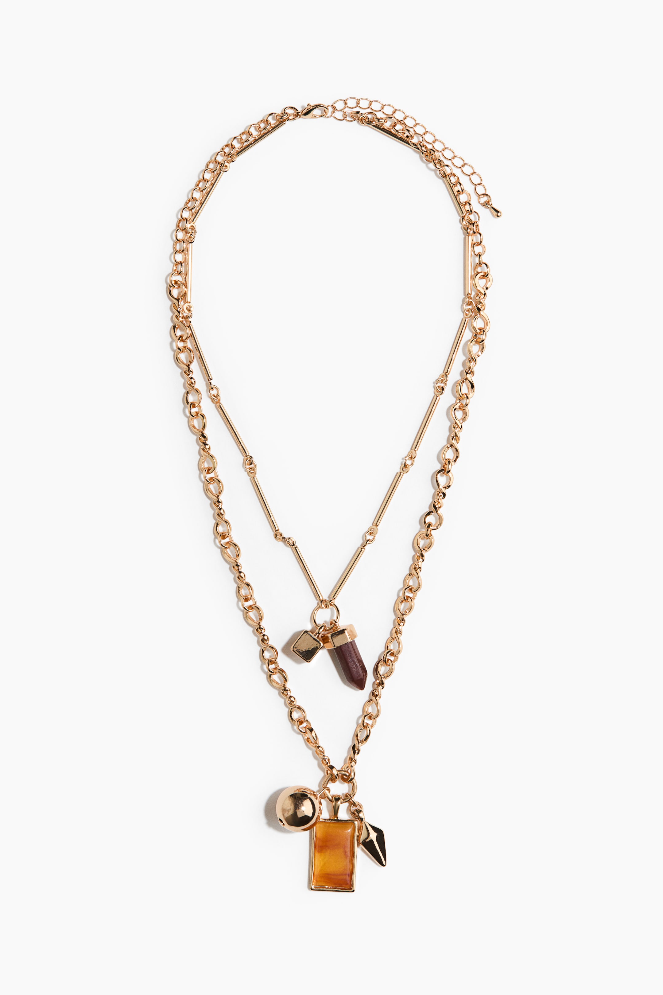 View larger image: A layered gold-tone necklace lies flat, showcasing two distinct chains. The upper chain features a faceted dark red crystal pendant and a small square charm, while the lower chain presents a round gold-tone bead, a rectangular amber-colored pendant, and a pointed gold-tone charm.