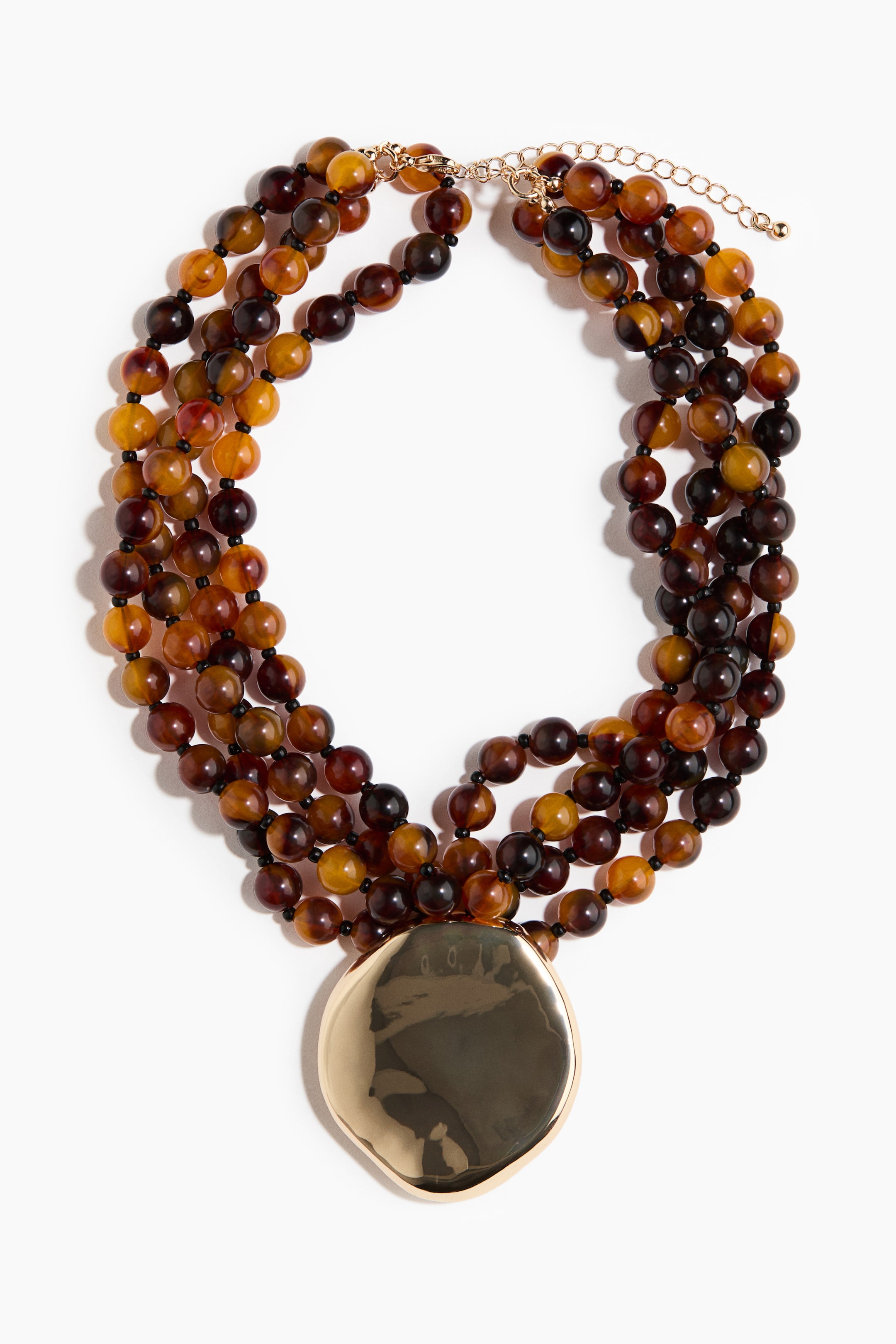 הצגת תמונה גדולה יותר: השרשרת כוללת חמישה גדילים בשכבות של חרוזים עגולים, שקופים בצבע ענברי וחומים אטומים, המשולבים עם חרוזי מפריד קטנים שחורים. תליון מתכתי גדול ובעל צורה בלתי סדירה, בגוון זהב, ממוקם במרכז הקדמי ומחזיר אור, והשרשרת נסגרת בסגר לובסטר בגוון זהב ובשרשרת מאריכה.