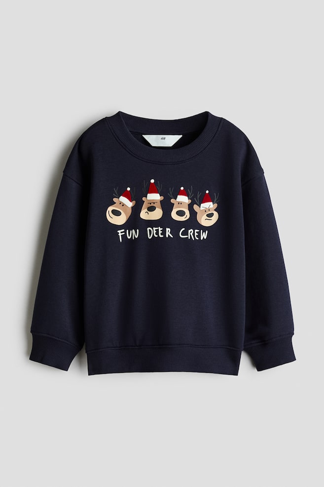 A navy blue sweatshirt is shown from the front, featuring a graphic print of four cartoon reindeer wearing red Santa hats above the white text "FUN DEER CREW". The garment is designed with a ribbed crew neckline, dropped shoulders, and ribbed cuffs and hem. - 1