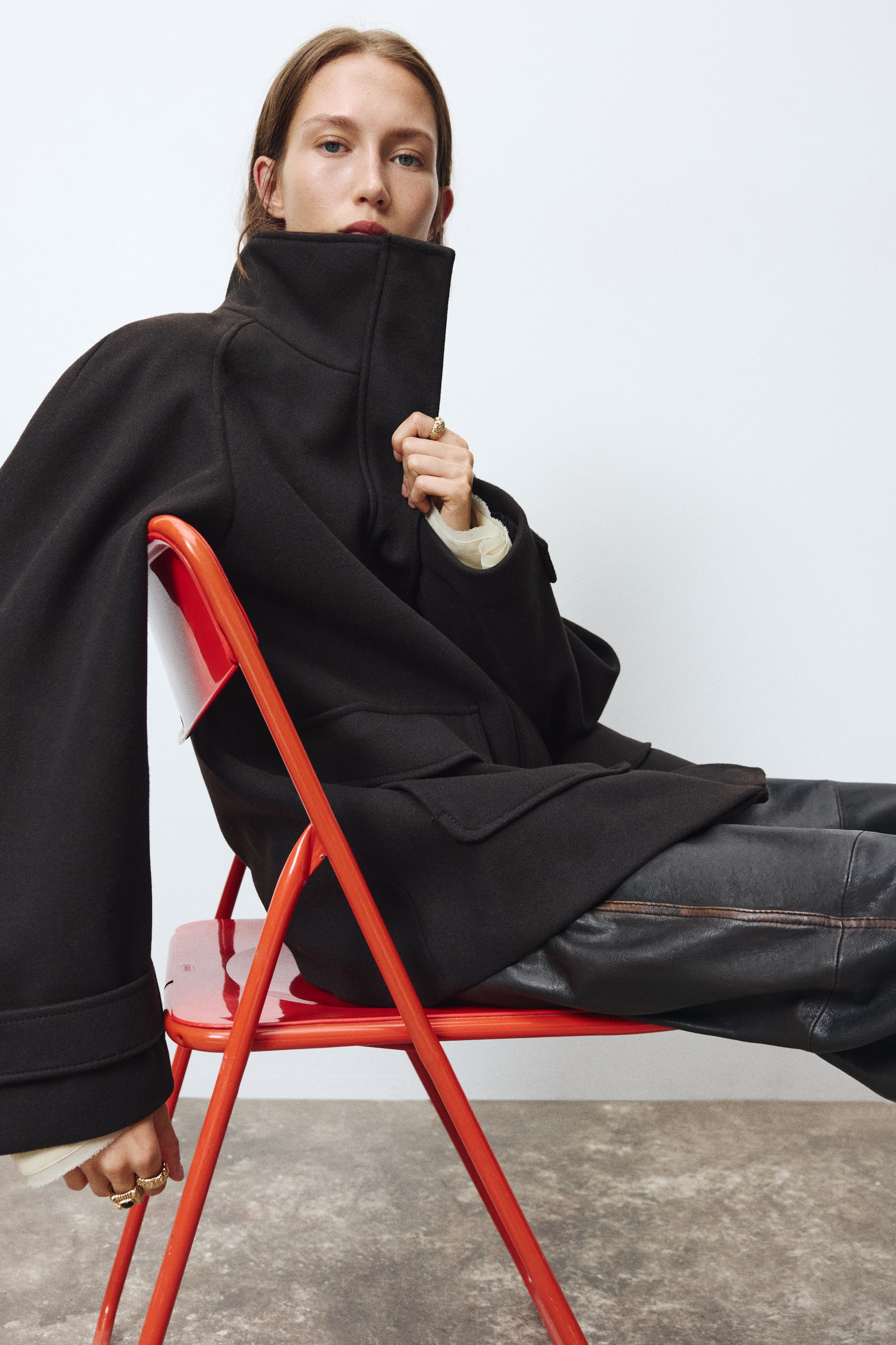 View larger image: A black felted fabric coat featuring a high funnel collar, raglan sleeves, and front patch pockets is being worn by the model seated on a red chair. The model is holding the upright collar close to their chin, with the coat styled over dark leather trousers.