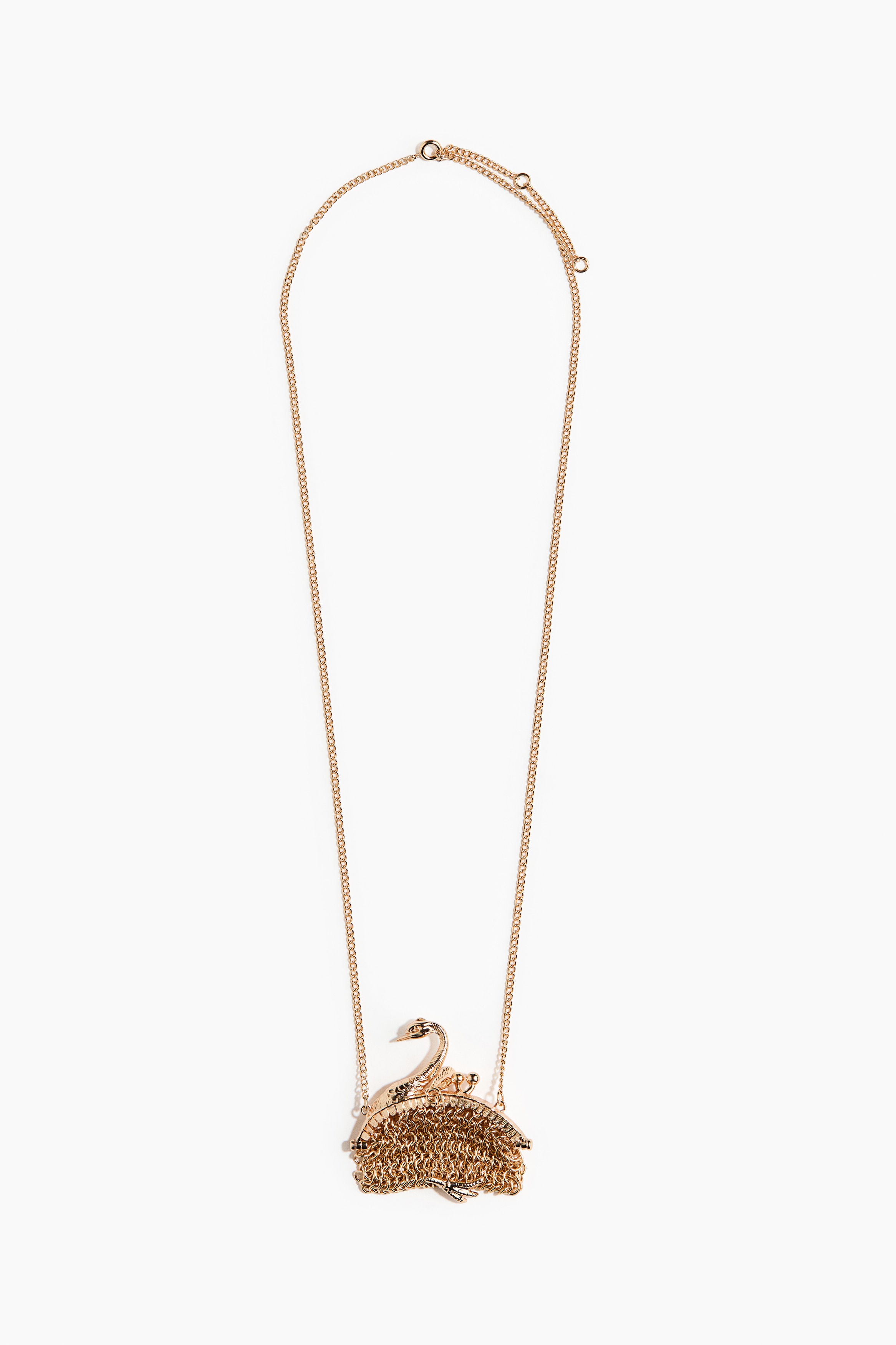 Een goudkleurige metalen ketting met een lange schakelketting en een gestructureerde hanger in de vorm van een klein tasje met zwanenmotief wordt gedragen door het model. De hanger rust tegen een zwarte kanten top, gedragen onder een open, gestructureerde zwarte jas.