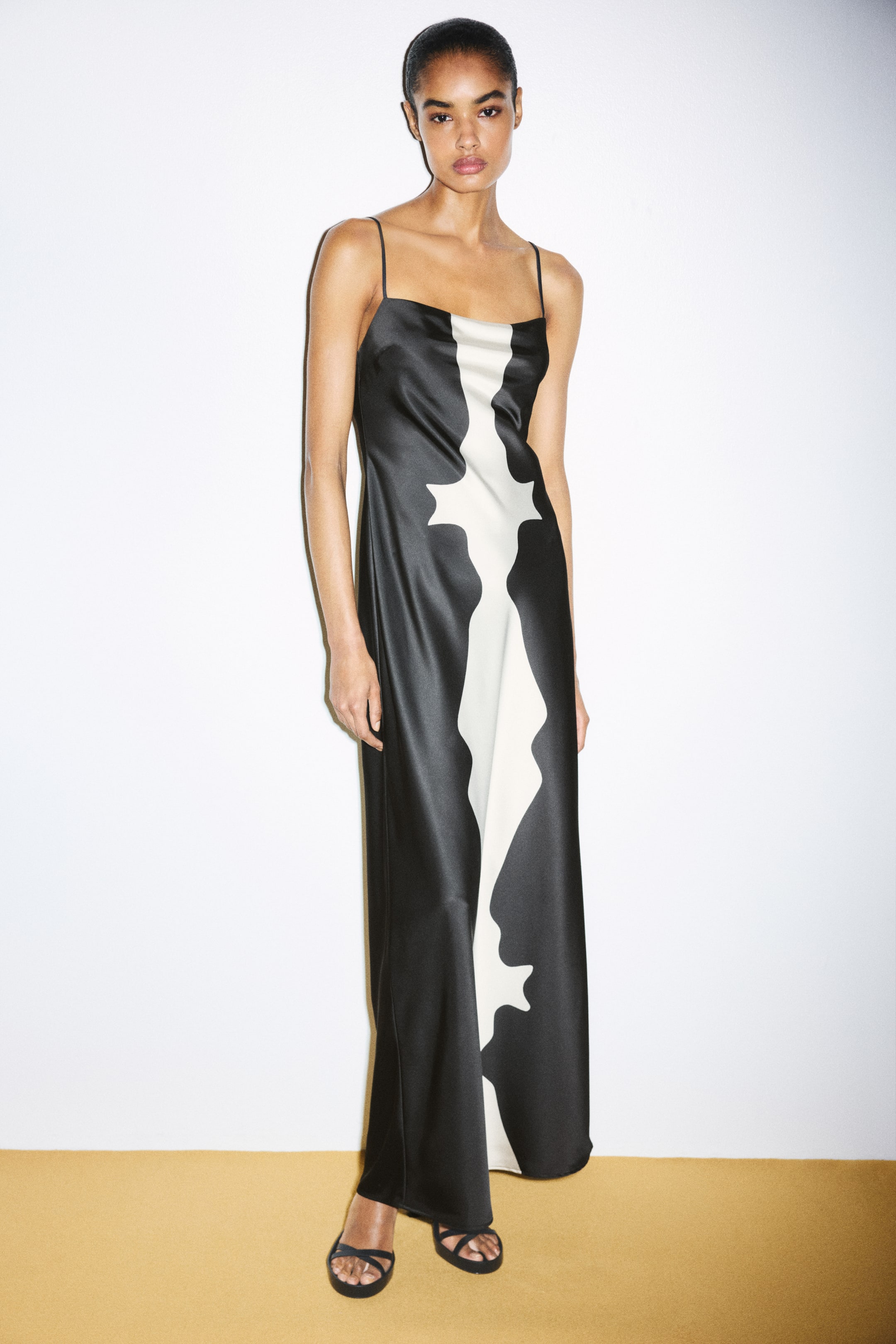 View larger image: A black satin maxi dress featuring a central white abstract motif is worn by the model. The dress is designed with a straight neckline and thin shoulder straps, and styled with black strappy sandals.