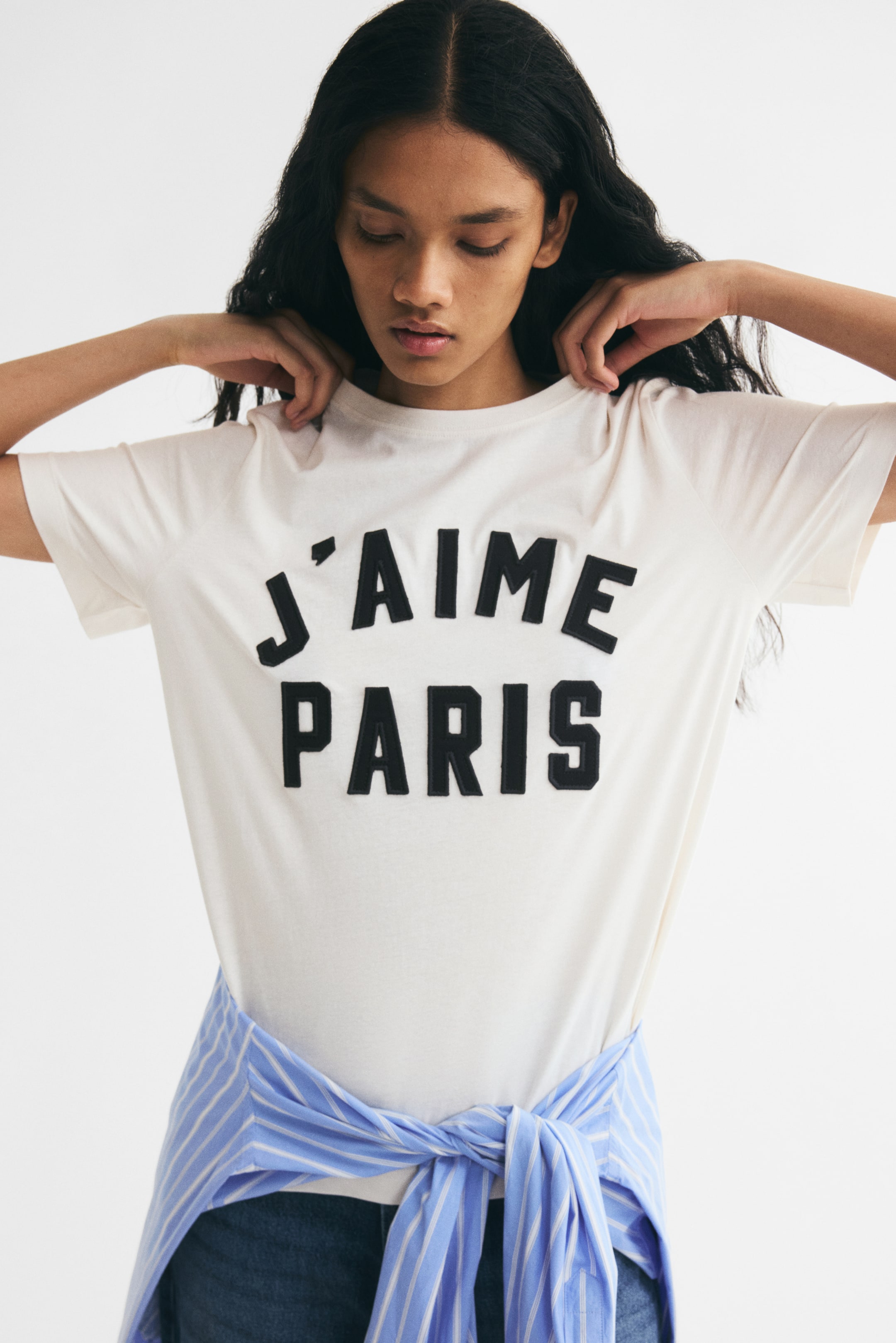 Xem ảnh lớn hơn: Một chiếc áo thun jersey cotton màu kem với dòng chữ "J'AIME PARIS" in đậm màu đen ngang ngực đang được người mẫu mặc, đồng thời điều chỉnh phần vai của trang phục bằng cả hai tay. Áo thun được phối cùng một chiếc áo sơ mi kẻ sọc xanh và trắng buộc quanh eo.