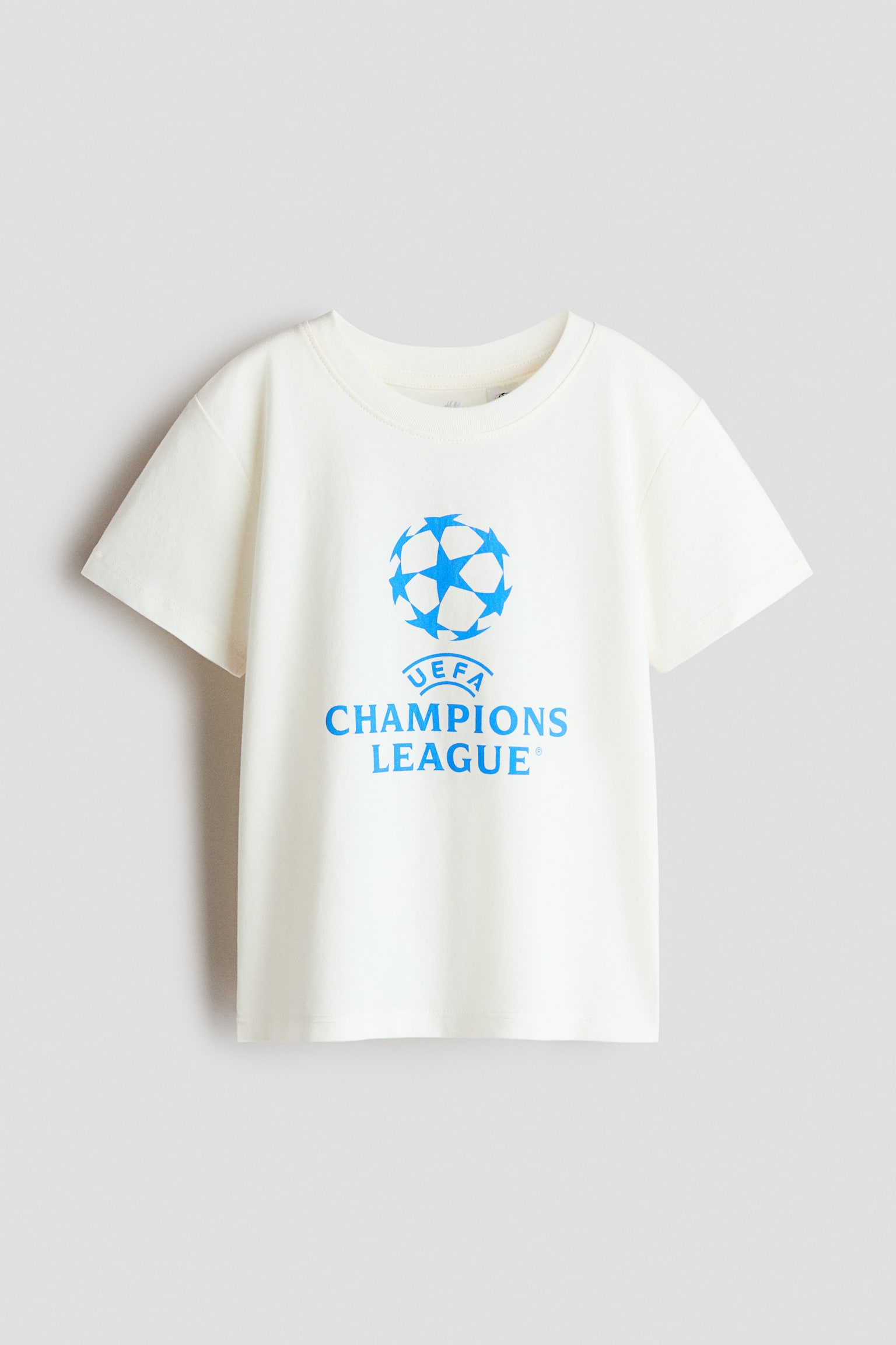 An off-white cotton jersey T-shirt is shown from the front, featuring a round rib-trimmed neckline and gently dropped shoulders. The front of the T-shirt displays a printed graphic of the bright blue UEFA Champions League starball logo and text.