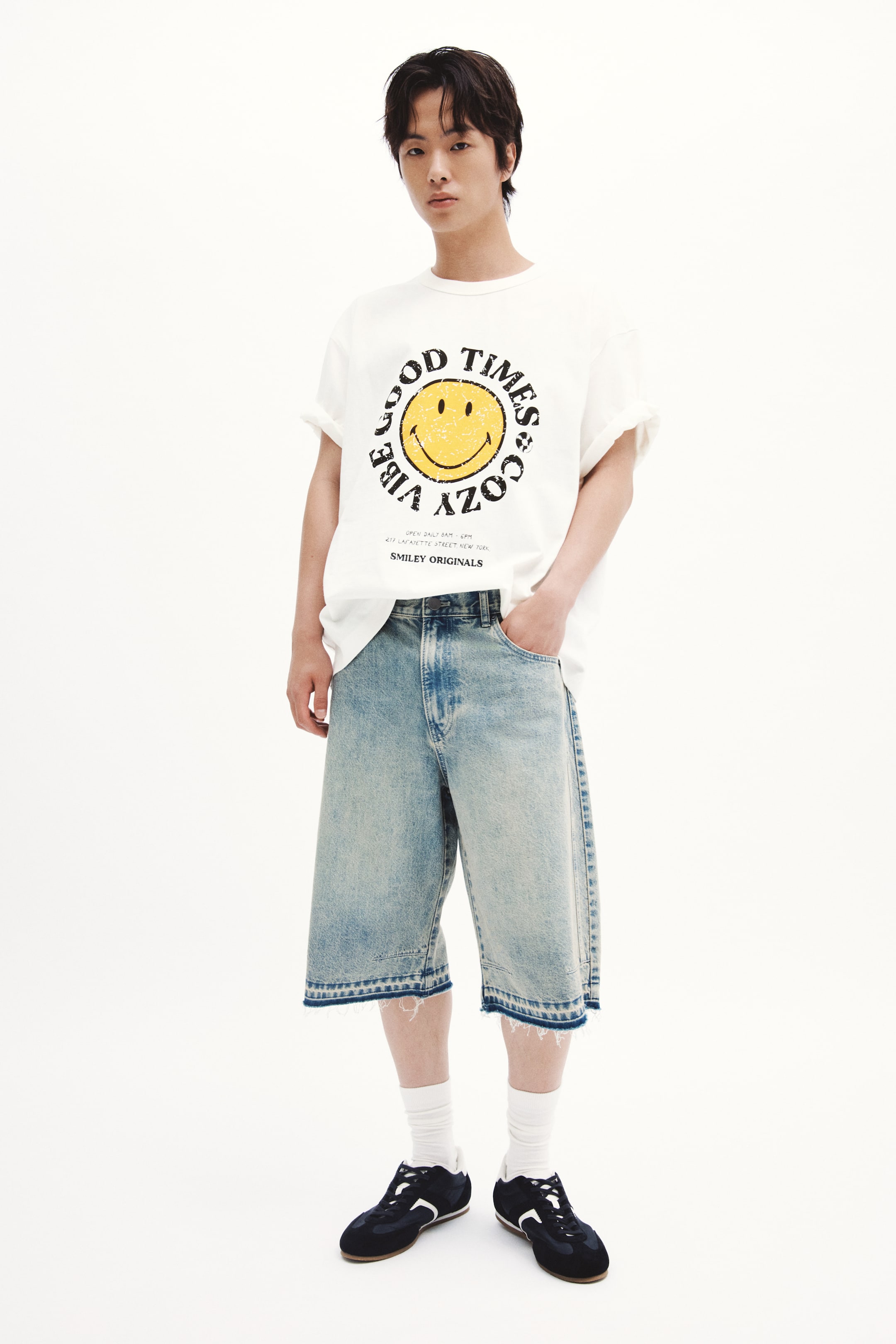 Xem ảnh lớn hơn: Một chiếc áo thun jersey cotton dày dặn có in hình được người mẫu mặc, với cổ tròn viền gân, vai rớt và dáng rộng rãi để tạo nên dáng vẻ thoải mái nhưng không quá rộng. Một đồ họa hình tròn với mặt cười màu vàng và dòng chữ "GOOD TIMES COZY VIBE" được hiển thị ở phía trước.