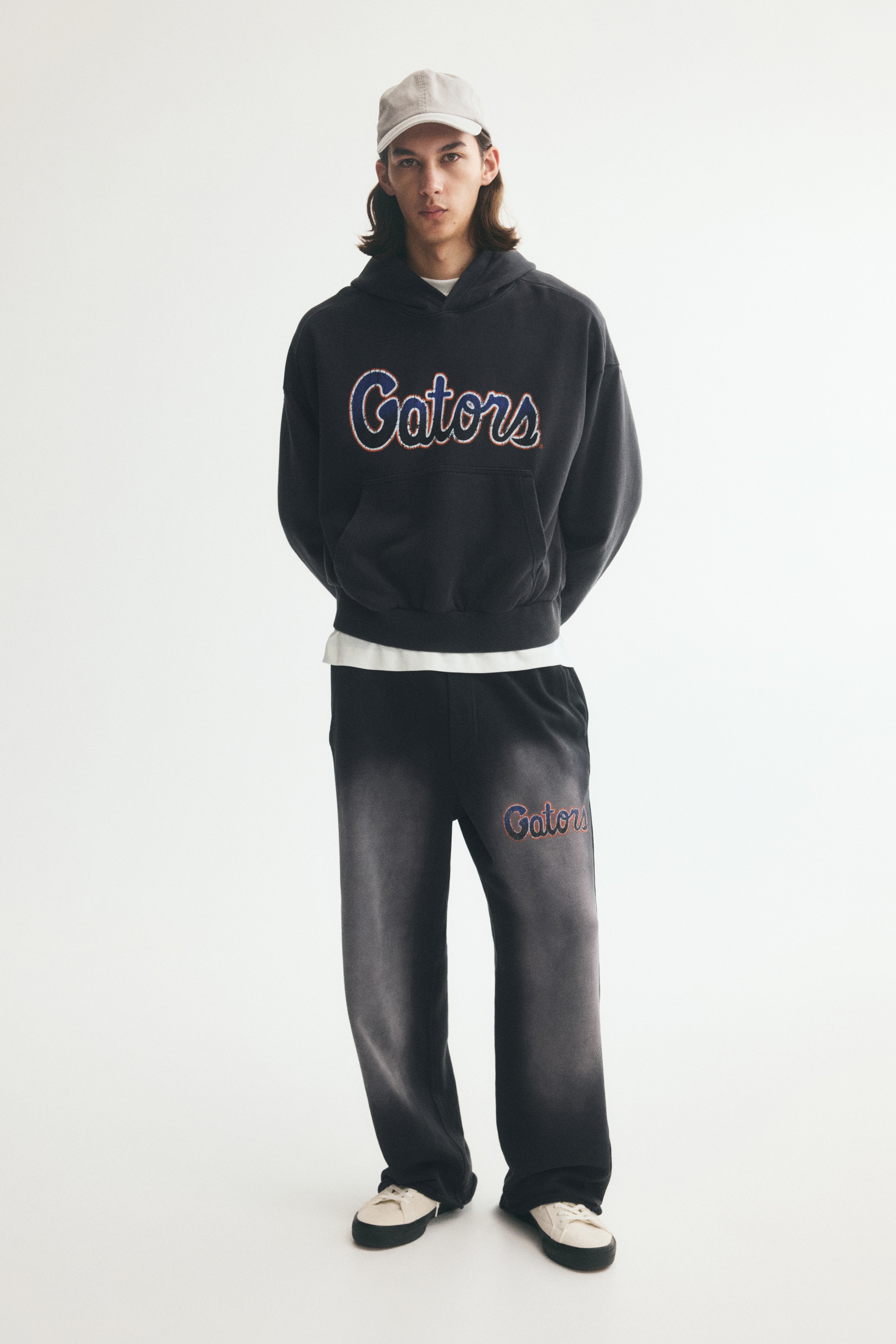 Black wide-leg sweatpants are shown from the front, featuring an elasticized waistband with a matching drawstring and side slip pockets. The pants display a prominent faded gray spray effect running vertically down the centre of each leg, with a blue cursive "Gators" logo outlined in orange on the left thigh.