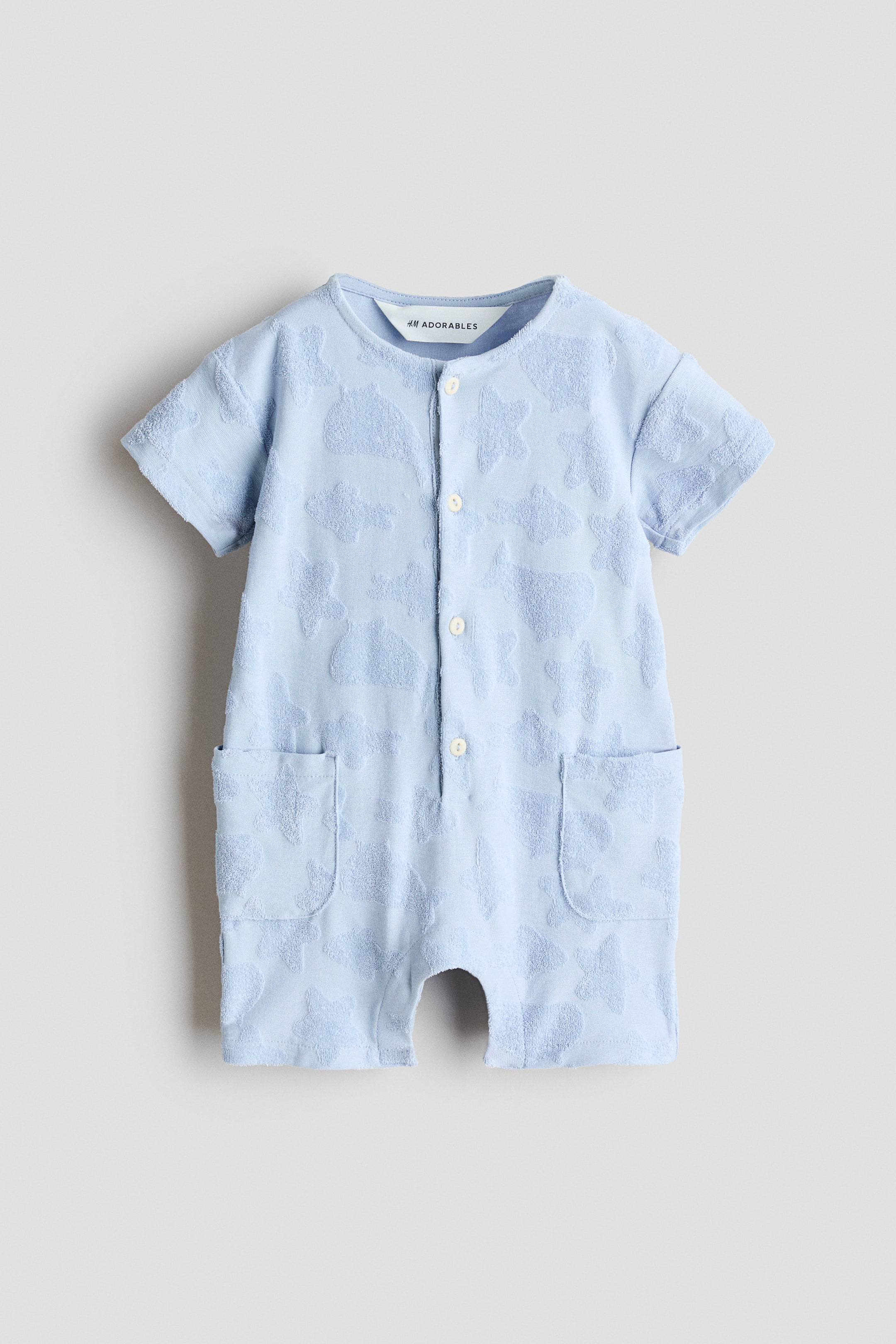 Ver imagen más grande: Un mono de manga corta en color azul claro de H&M Adorables se muestra de frente, confeccionado en un tejido de jersey texturizado con un patrón en relieve. Está diseñado con un escote redondo, una tapeta de botones en la parte delantera y dos bolsillos de parche en la parte inferior delantera.
