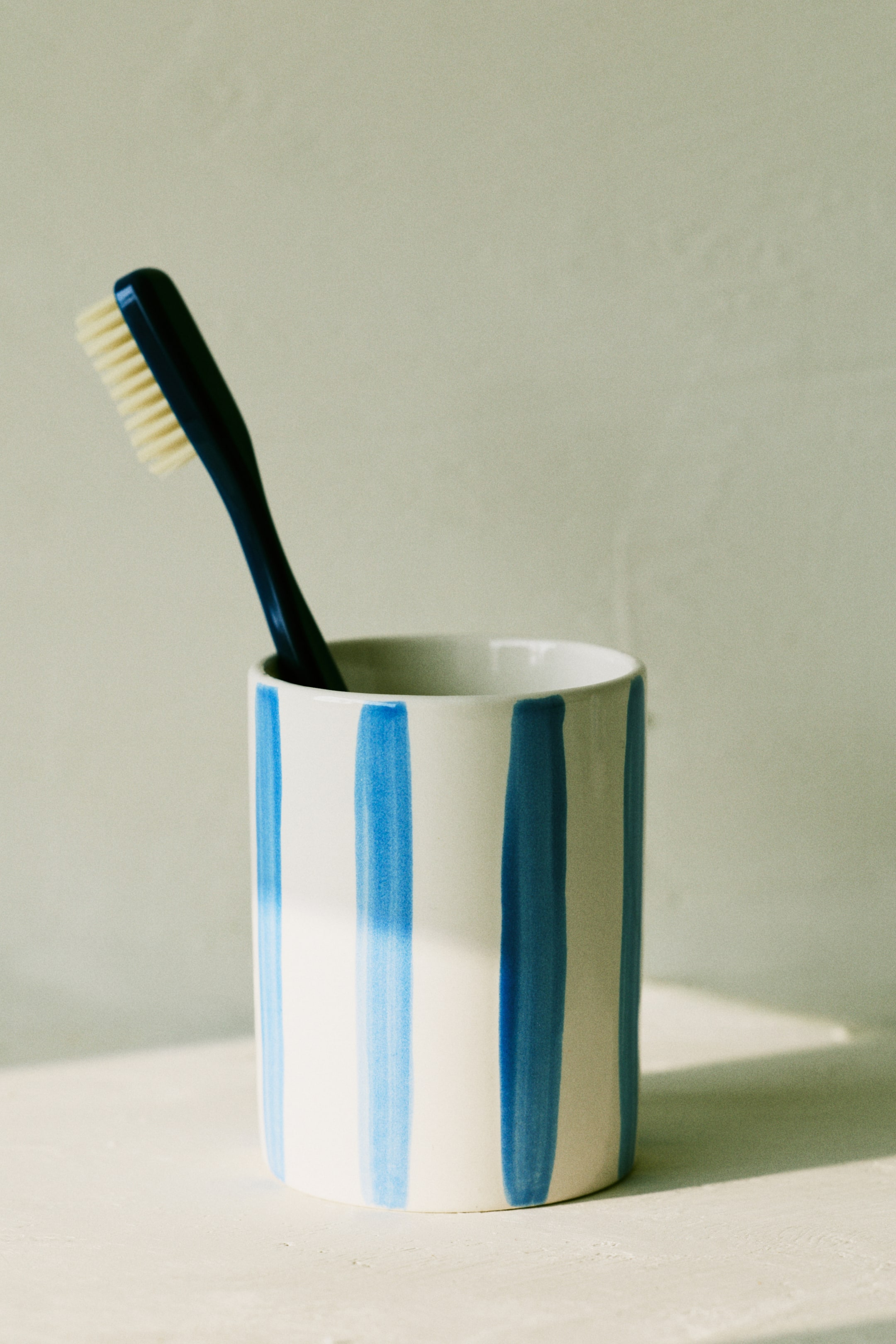 View larger image: A stoneware toothbrush mug features a white exterior with vertical blue stripes, shown from the front. A dark blue toothbrush with light bristles rests inside.