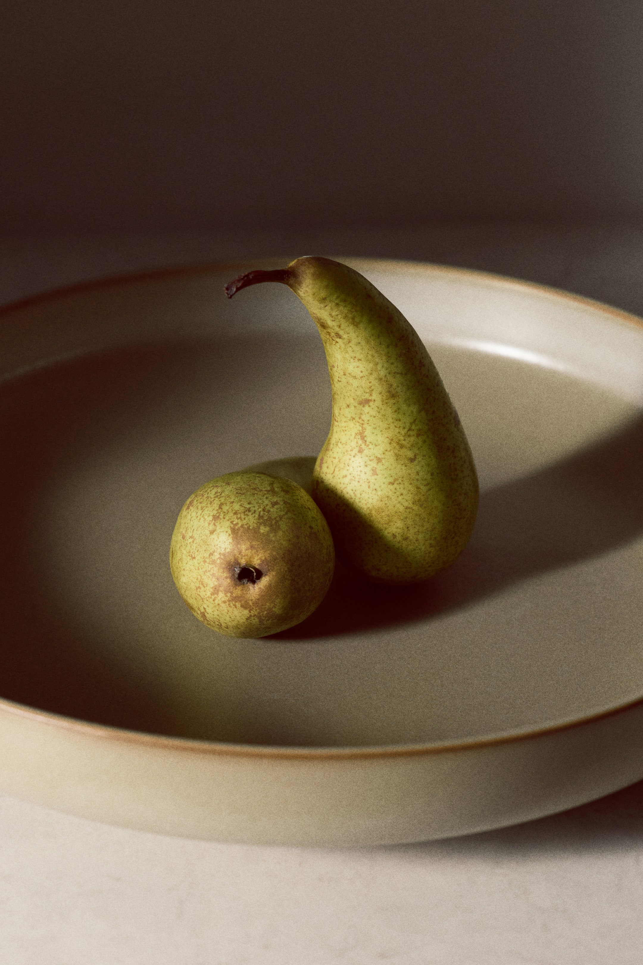 View larger image: Two green-yellow pears are resting in a shallow, light olive-green bowl. The larger, elongated pear is standing upright, while the smaller, rounder pear is lying flat beside it, both displaying brown speckles on their skin.