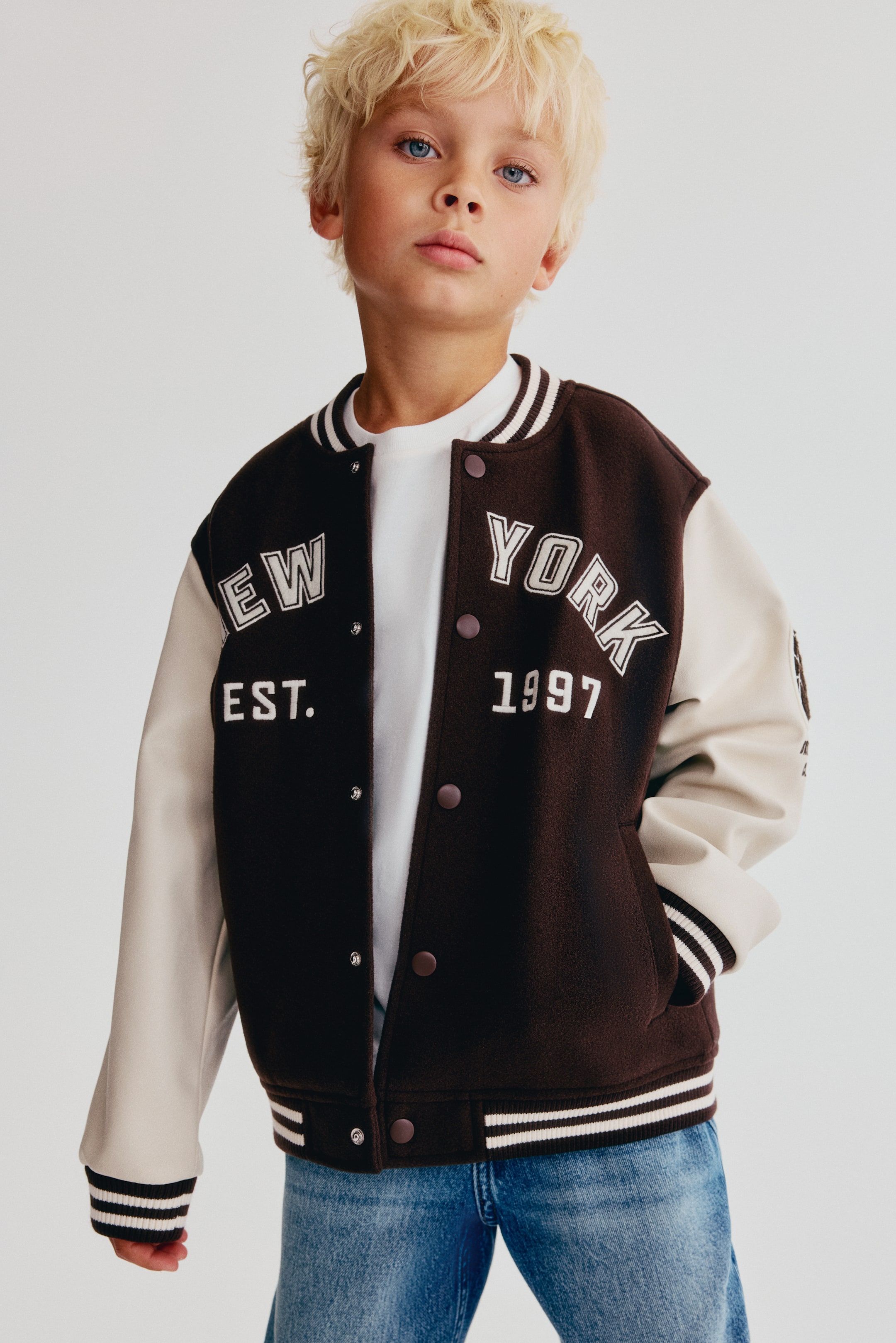 Grotere afbeelding bekijken: Een van voren afgebeelde baseballjack met een donkerbruine gevilte body met contrasterende crèmekleurige mouwen en de tekst "NEW YORK EST. 1997" op de borst. Het jack is ontworpen met een geribde kraag, manchetten en zoom met strepen, drukknoopsluiting aan de voorkant en paspelzakken aan de zijkanten, open gestyled over een wit T-shirt.