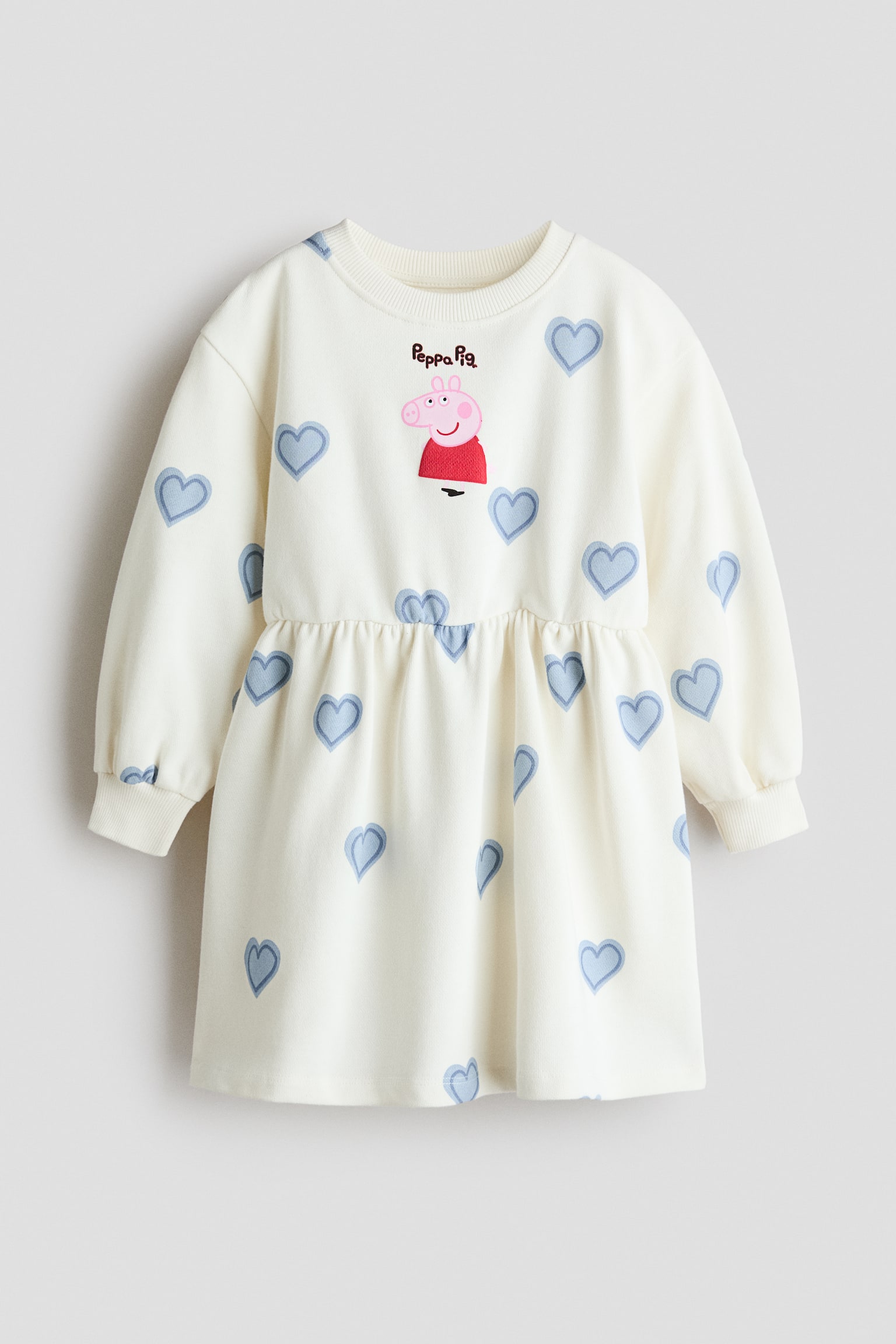 A cream-coloured fleece sweatshirt dress is shown from the front, featuring a round, rib-trimmed neckline, dropped shoulders and long balloon sleeves with wide ribbed cuffs. The dress is patterned with blue heart outlines and features a central Peppa Pig graphic on the chest above a gathered waist and a flared skirt.