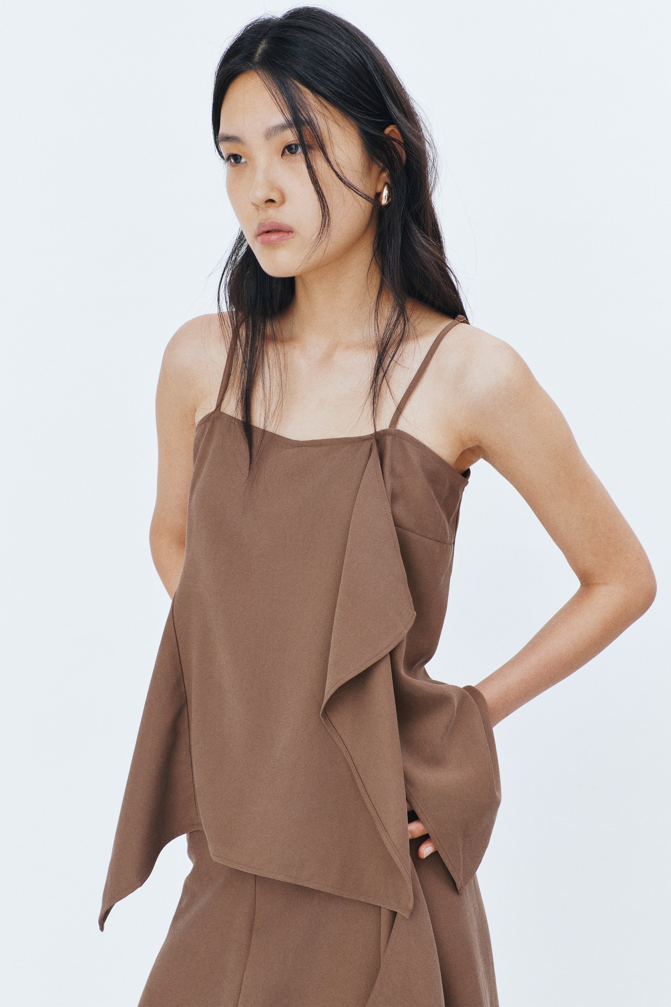 View larger image: Model tersebut memakai baju atasan berwarna coklat dengan potongan longgar, menampilkan garis leher lurus dan tali bahu yang sempit serta boleh laras. Baju atasan ini memperlihatkan rekaan lilit hadapan yang dijahit dengan lipatan beralun pada satu sisi, manakala satu flounce lebar melengkung di pinggang pada sisi bertentangan, menghasilkan kelim asimetrik di mana tangan model itu sedang berehat.