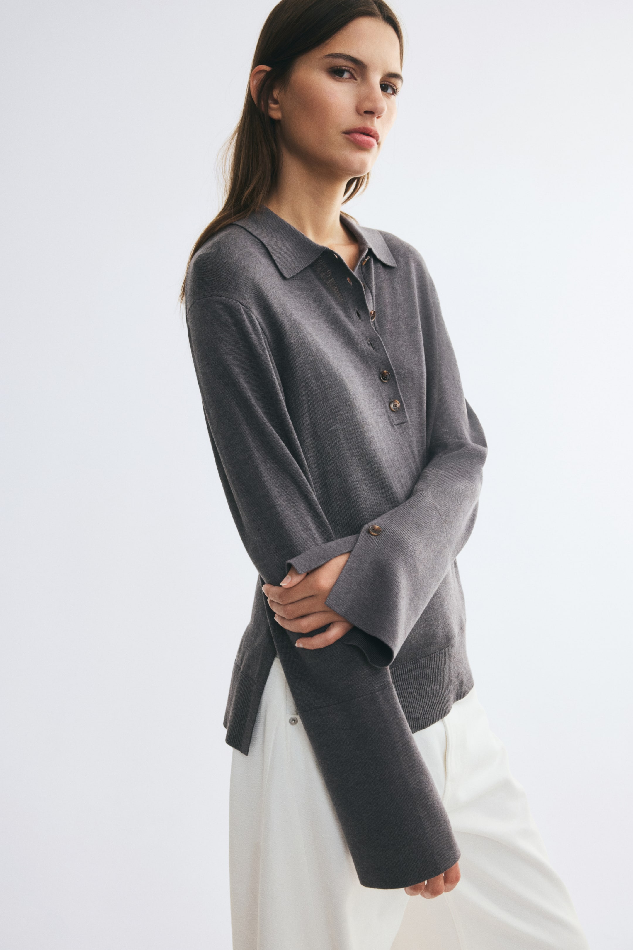 View larger image: A grey, fine-knit polo shirt is worn by the model, featuring a ribbed collar, a button placket, and dropped shoulders. The shirt has long sleeves with wide buttoned cuffs and slits, which the model is holding with one hand while wearing white trousers.