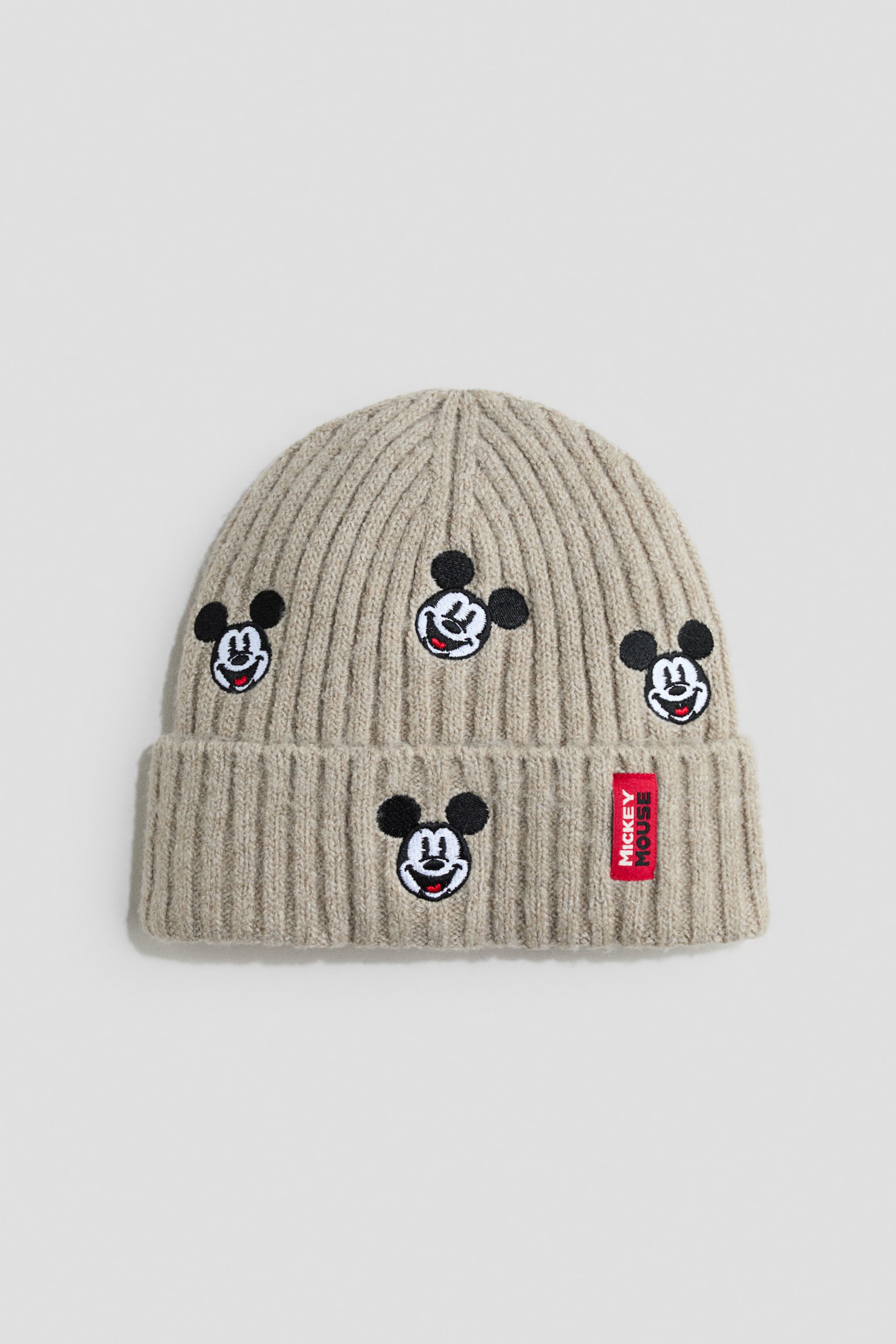 A beige ribbed-knit beanie is shown front-facing, featuring a folded brim and four embroidered Mickey Mouse face patches scattered across the crown and brim. A small, vertical red label with white âMICKEY MOUSEâ lettering is attached to the right side of the folded brim.