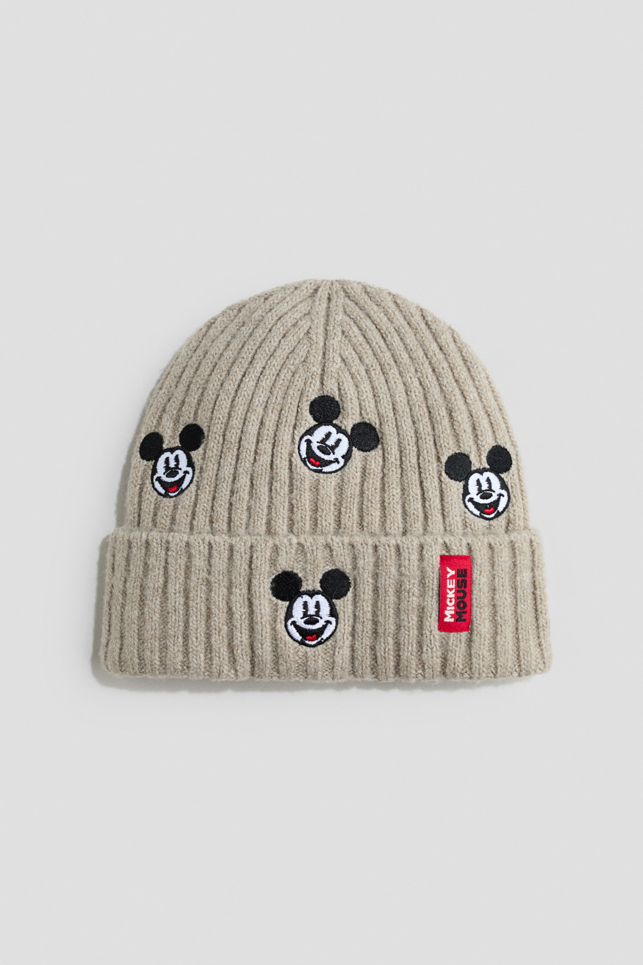 View larger image: A beige ribbed-knit beanie is shown front-facing, featuring a folded brim and four embroidered Mickey Mouse face patches scattered across the crown and brim. A small, vertical red label with white “MICKEY MOUSE” lettering is attached to the right side of the folded brim.