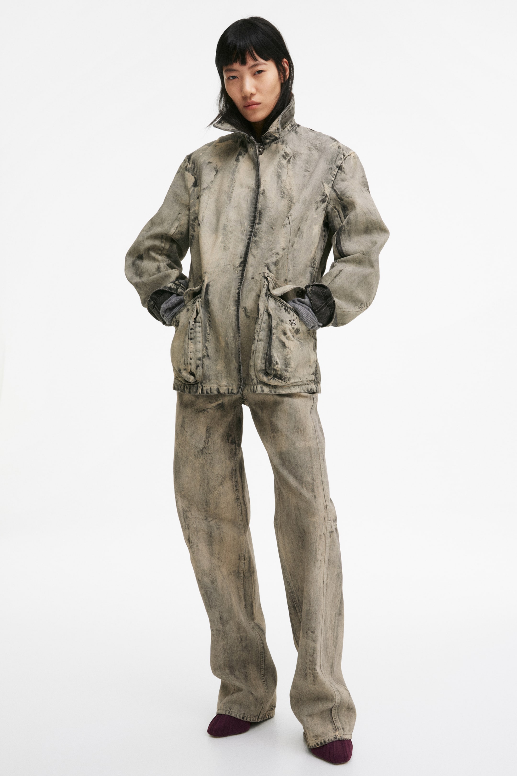 View larger image: The model is wearing a jacket in rigid cotton denim, featuring a coated, worn appearance, a pointed collar, a front placket with concealed press-studs, and a straight-cut hem. The jacket also includes two bellows front pockets with flaps, and the model is placing their hands into these pockets, revealing grey ribbed cuffs.