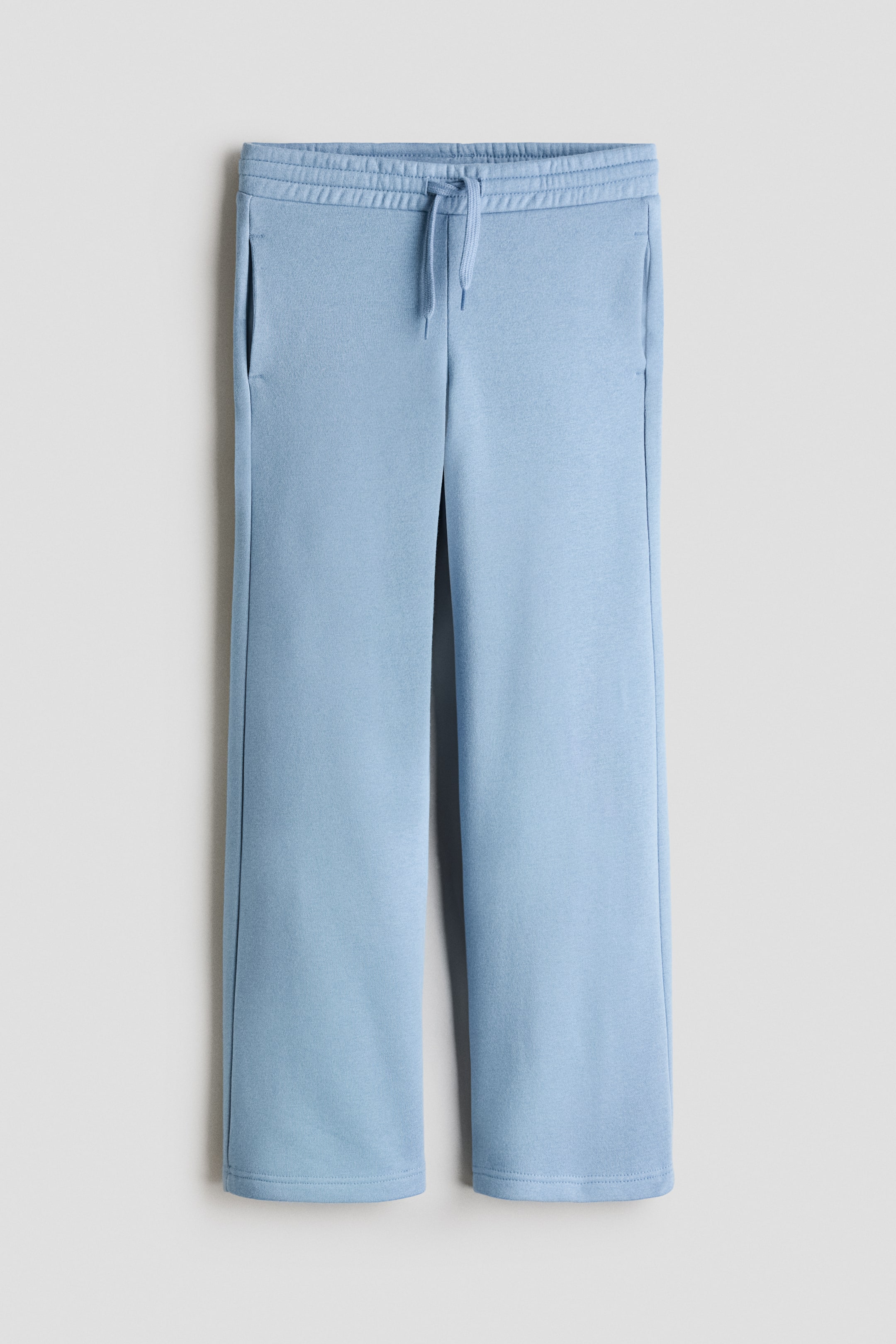 View larger image: Light blue joggers are laid flat, showcasing a smooth, sweatshirt-style fabric. They feature an elasticated waist with a tied drawstring, discreet side pockets, and straight legs with simple hems.
