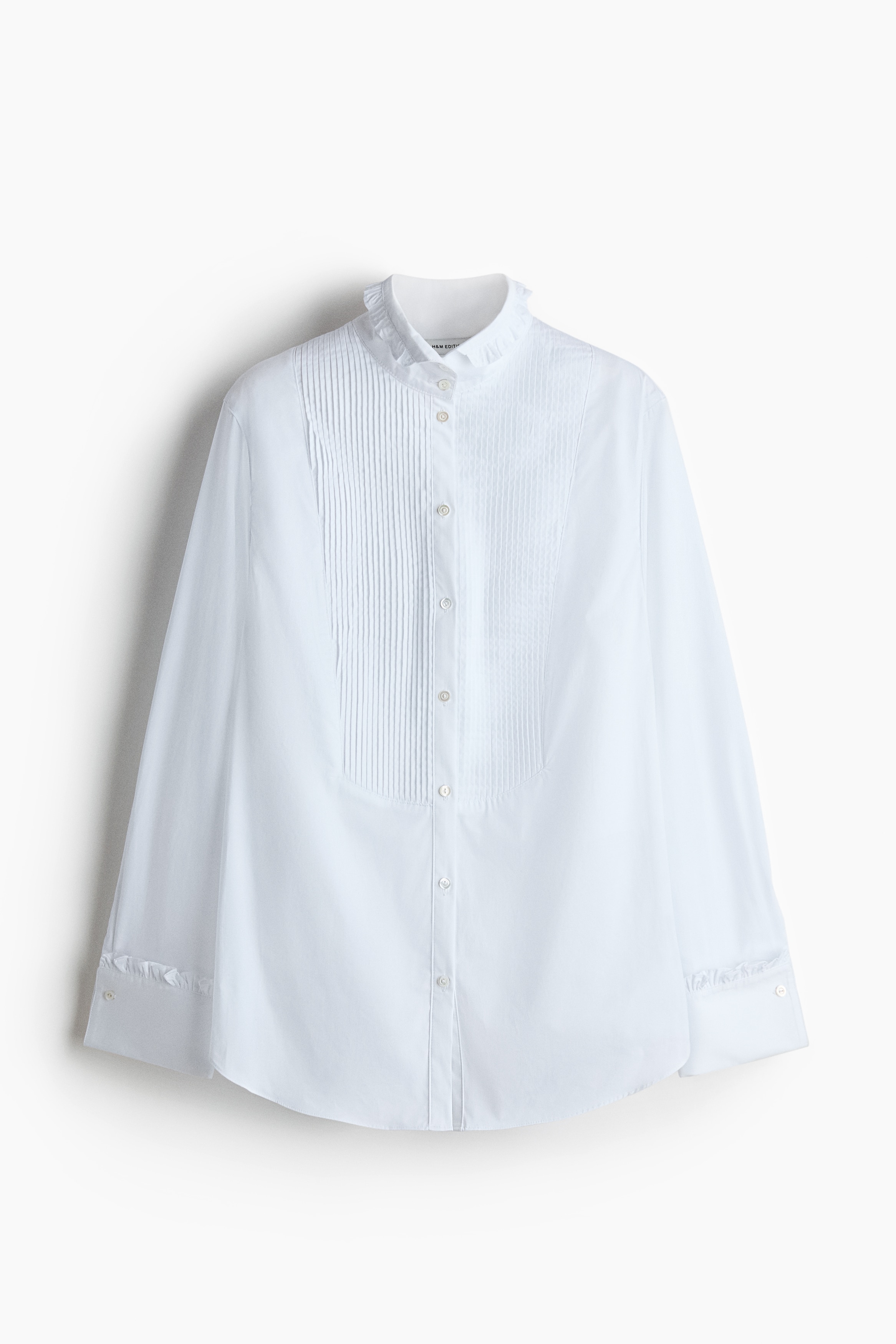 La modelo lleva una camisa blanca de algodÃ³n con pechera plisada, cuello mao con ribete de volantes y puÃ±os con volante. La modelo sostiene un bolso de piel marrÃ³n texturizada y tiene una mano metida en el bolsillo de un pantalÃ³n oscuro.