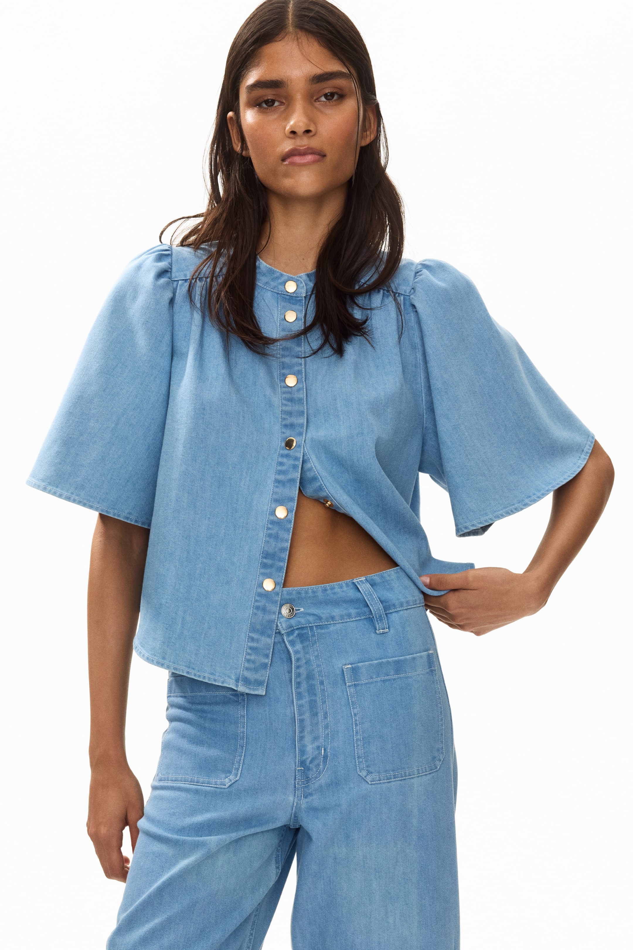 View larger image: The model is wearing light blue denim jeans that showcase a high waist and button closure, complete with front patch pockets. These wide-leg jeans boast a generous cut from the thigh down to the hem, with the model holding onto the waistband.