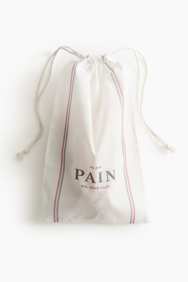 A light cream fabric drawstring bag is standing upright, facing forward. It features vertical red and grey stripes on either side of the central text 'un bon PAIN s'il vous plaît', where 'PAIN' is in a larger font, and a light-coloured cord drawstring is cinching its top. - 1