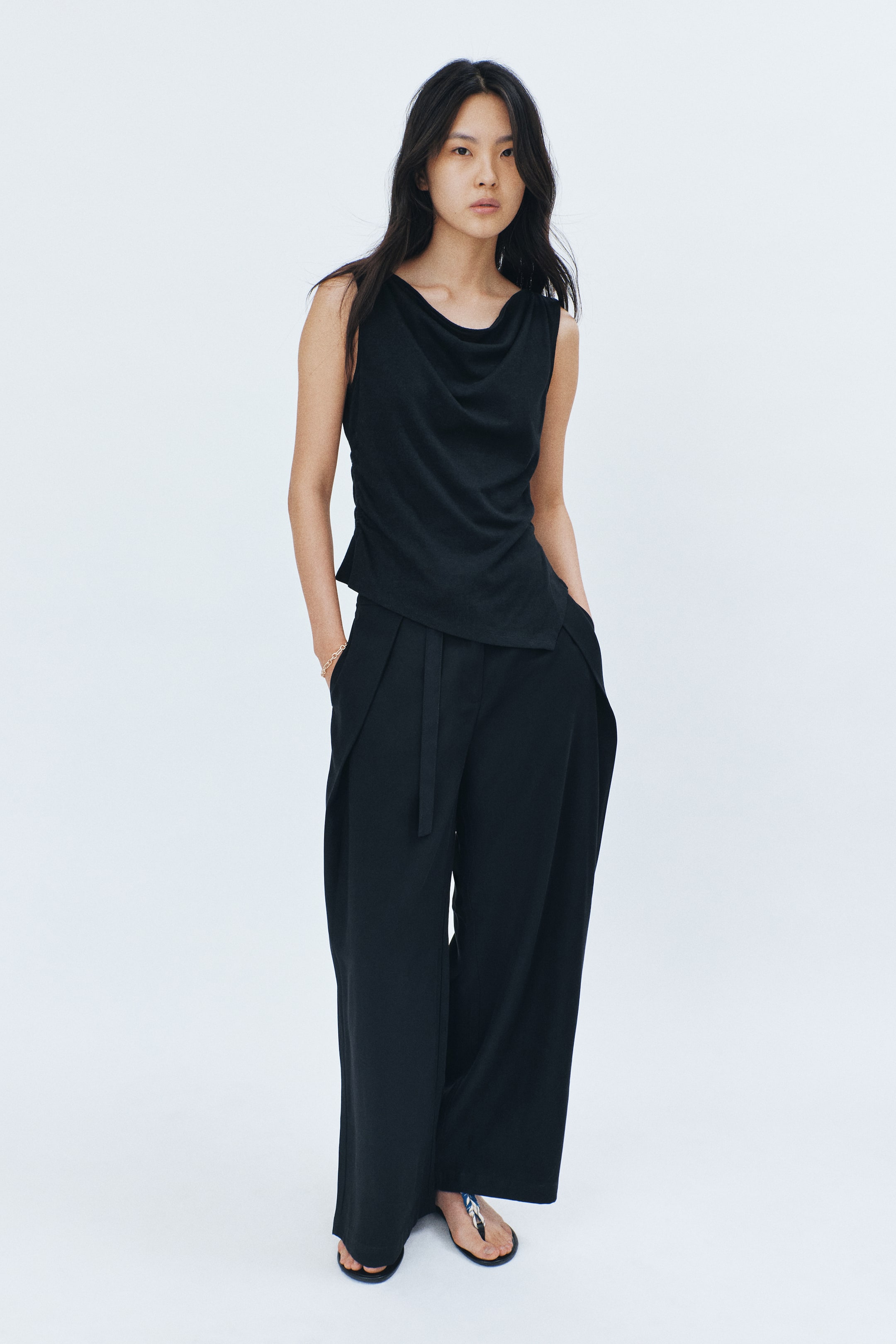 View larger image: A black sleeveless top with a cowl neck and an asymmetric hem is worn. The model pairs it with black wide-leg pants featuring front pleats and a tie belt, with her hands casually placed in the pants' pockets.