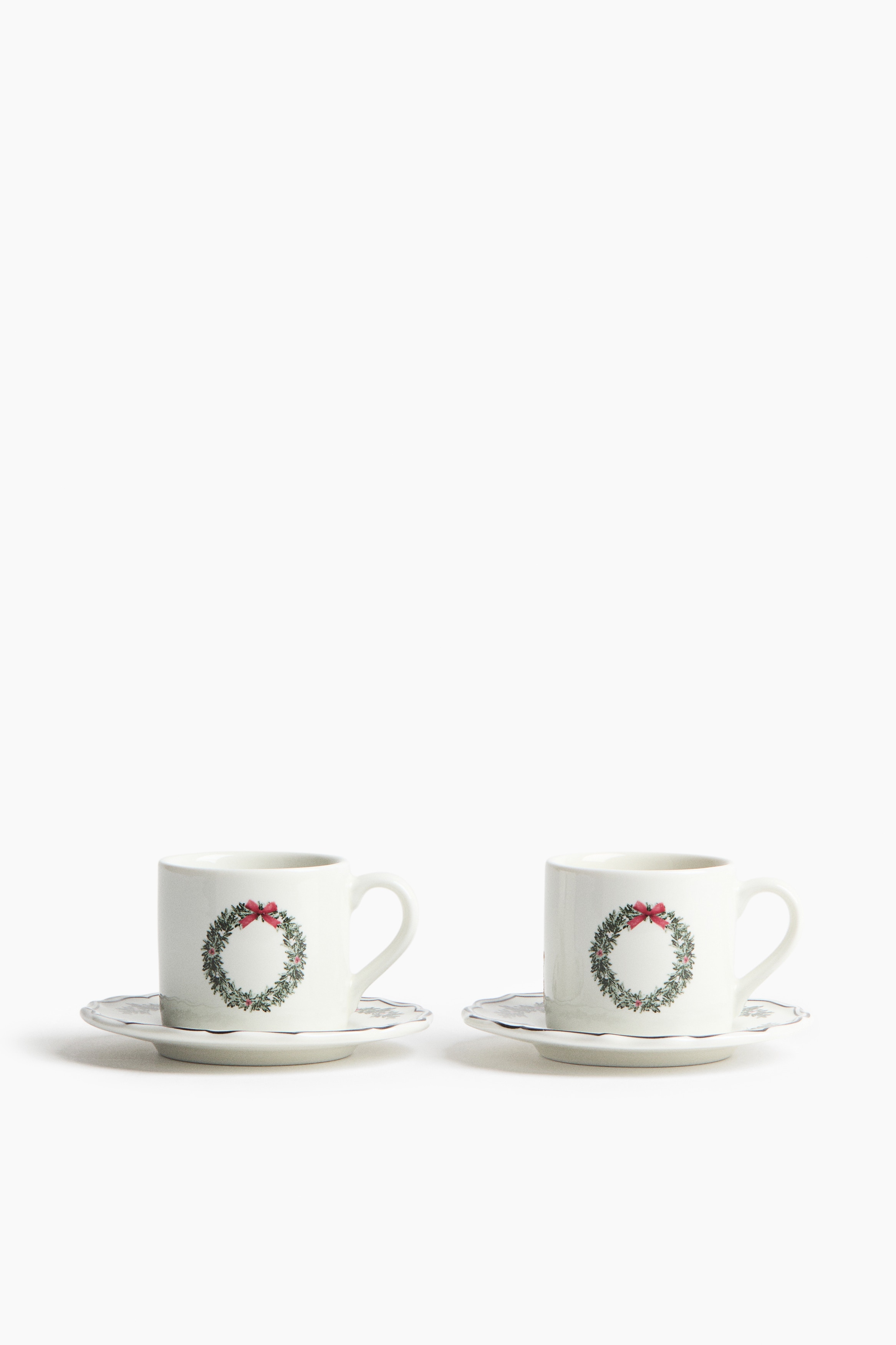 Une tasse Ã  espresso en cÃ©ramique avec un imprimÃ© festif de couronne verte et un nÅud rouge Ã  lâavant repose sur une sous-tasse assortie. La sous-tasse est dÃ©corÃ©e de motifs feuillus imprimÃ©s et prÃ©sente un rebord ondulÃ© de couleur dâaccent vert foncÃ©.