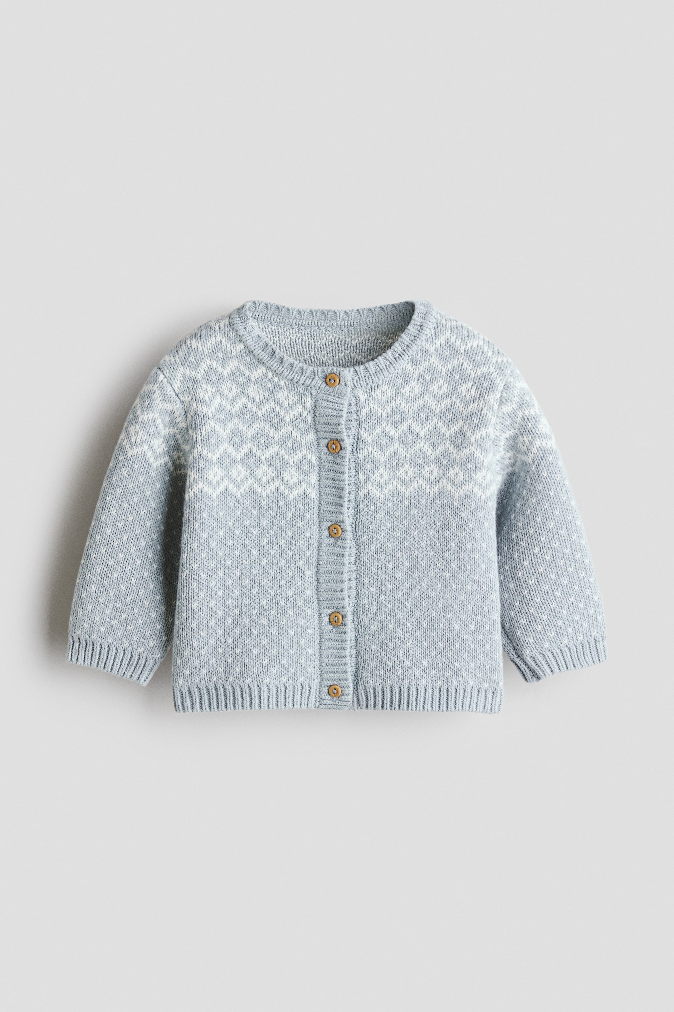 Xem ảnh lớn hơn: Một chiếc áo cardigan len màu xanh nhạt được chụp từ phía trước, với cổ tròn, năm nút cài màu nâu dọc giữa thân và tay dài raglan. Sản phẩm nổi bật với họa tiết dệt kim hình học màu trắng ở ngang ngực, điểm những chấm trắng nhỏ khắp bề mặt, hoàn thiện bằng cổ áo, cổ tay áo và gấu áo bo gân.