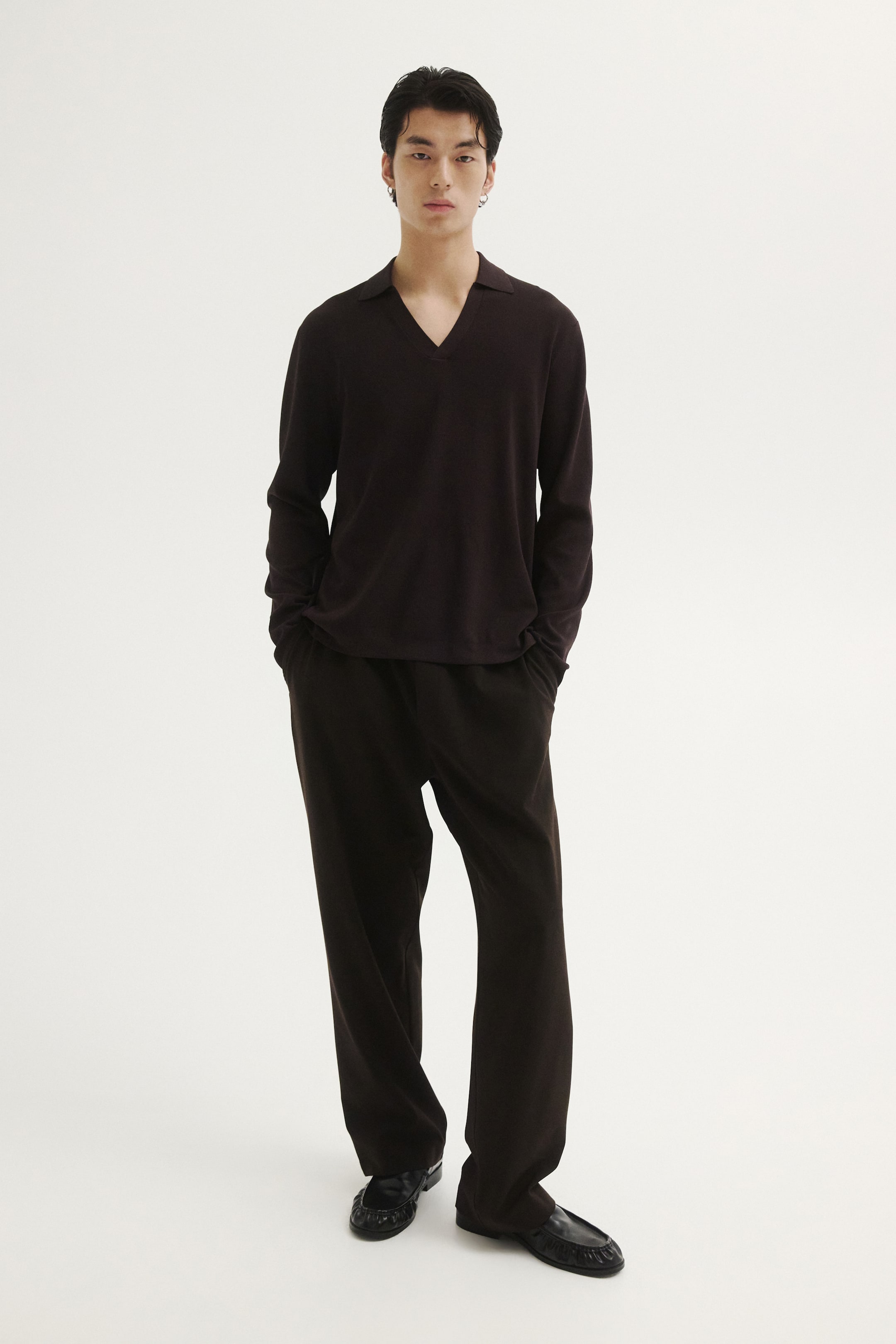 Ampliar la imagen: El modelo lleva puesto un polo de tejido de manga larga en color café oscuro, con cuello y escote en V. La prenda de corte relajado se combina con pantalones oscuros a juego y mocasines de piel negros.