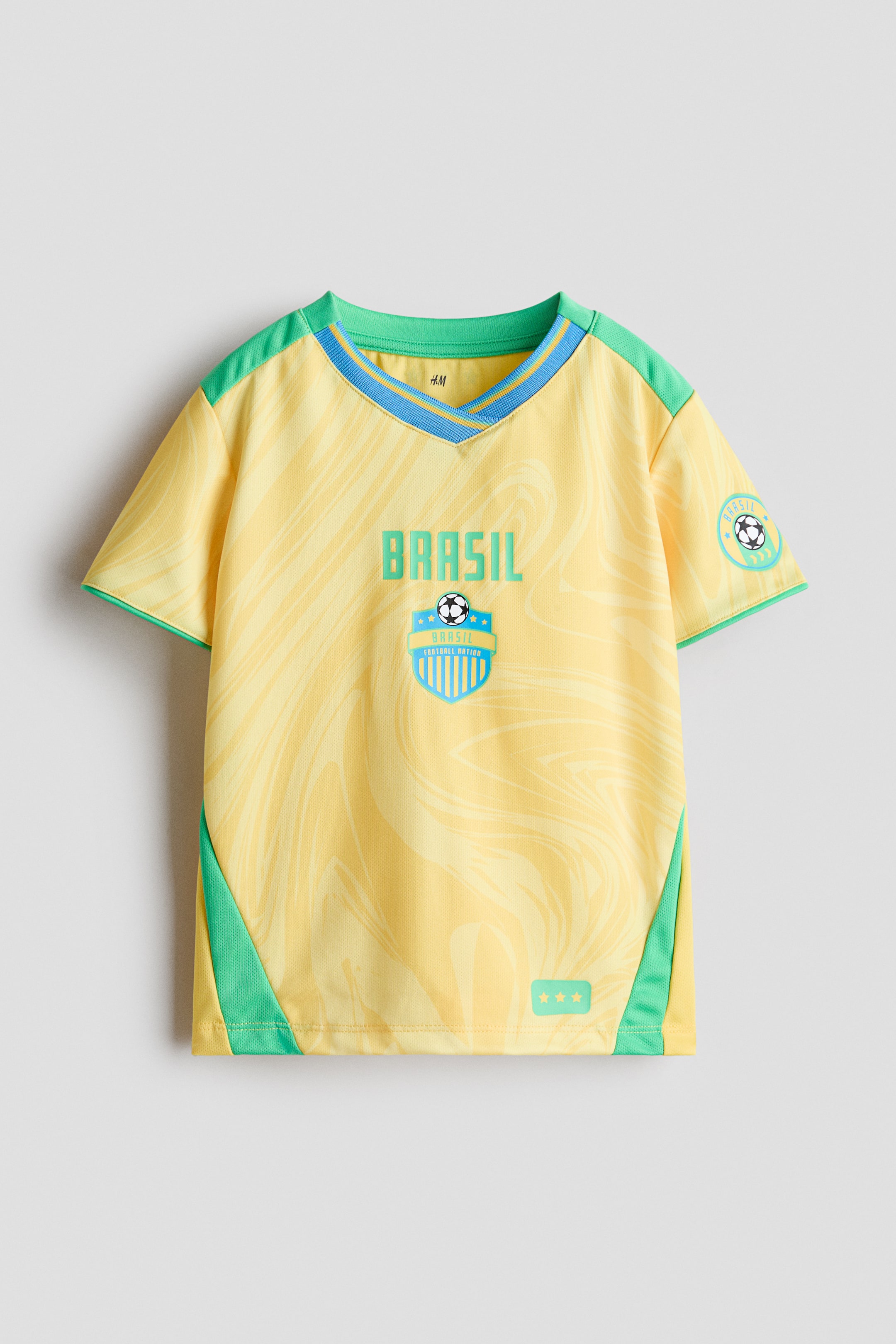 Ampliar la imagen: Una camiseta de fútbol de manga corta en malla amarilla impresa se presenta de frente, con un sutil patrón en espiral en toda su superficie. Muestra un escote en V con ribete, acentuado con detalles en verde y azul, paneles laterales de malla verde, y ribetes verdes en los puños de las mangas, junto con el texto "BRASIL", un emblema de escudo y tres símbolos de estrellas en el frente, además de un emblema circular en la manga derecha.