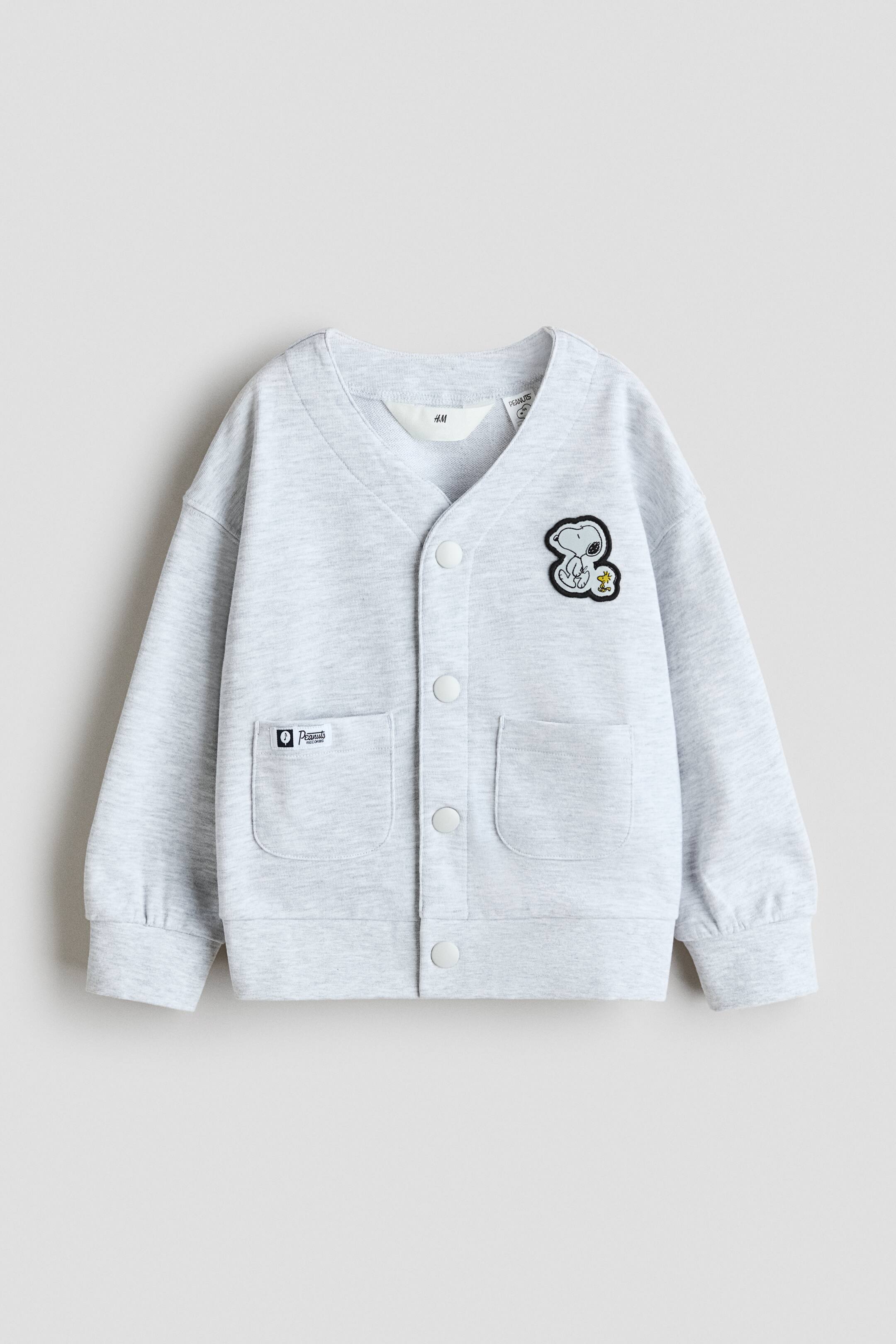 이미지 확대 보기: 정면을 향한 연한 그레이 멜란지 아동용 카디건으로, V넥과 다섯 개의 흰색 스냅 버튼 전면 여밈이 특징입니다. 패치 포켓이 두 개 보이며 오른쪽 가슴에는 Snoopy와 Woodstock 패치가 있고 왼쪽 포켓에는 작은 "Peanuts" 라벨이 부착되어 있습니다. 소매는 길고 소맷단이 셔링 처리되어 있으며 밑단은 골지 처리되어 있습니다.