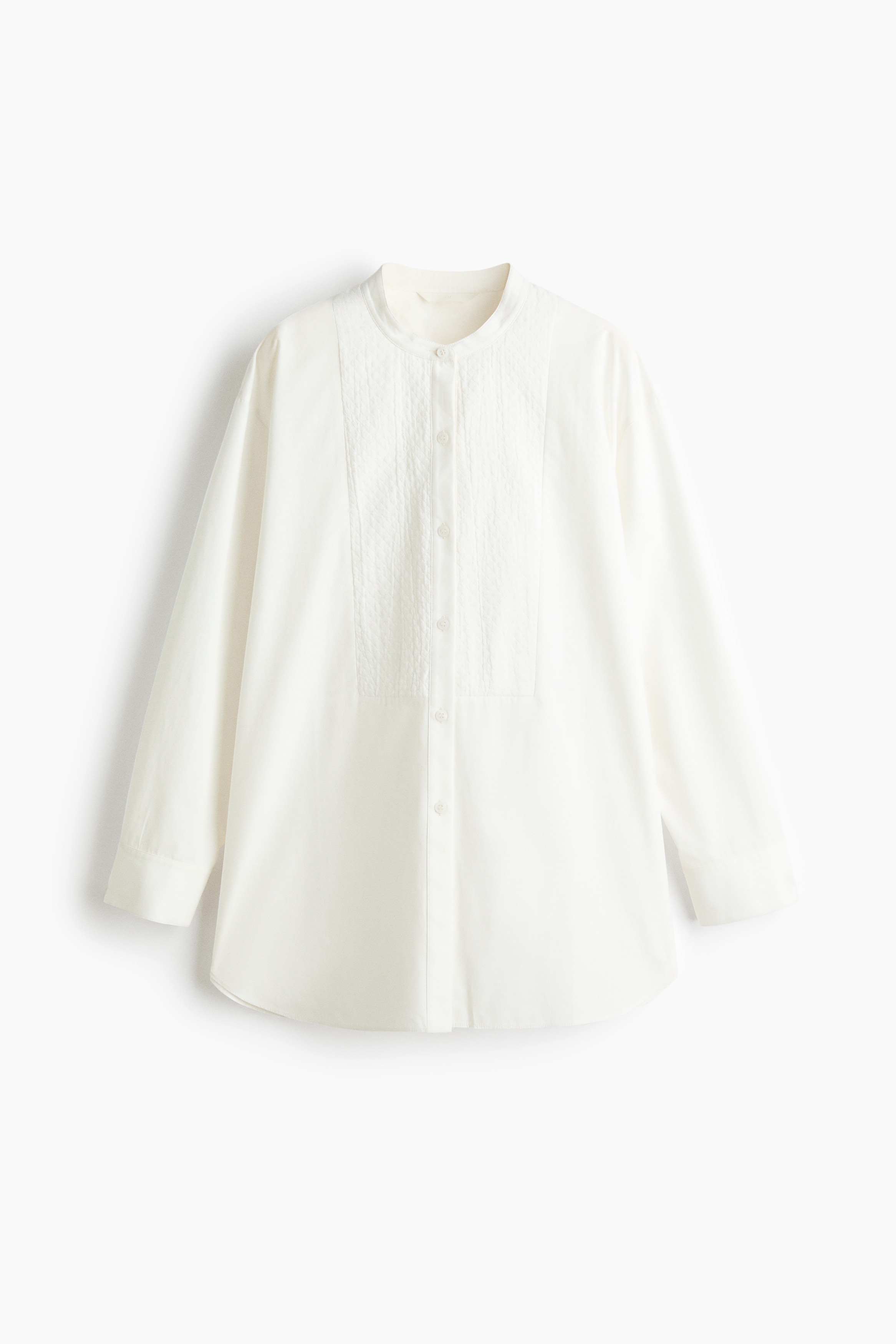 A white cotton-weave shirt featuring a band collar, a textured bib front, and dropped shoulders is worn by the model. The shirt is styled partially tucked into dark jeans, showcasing its long sleeves with button cuffs and relaxed fit.