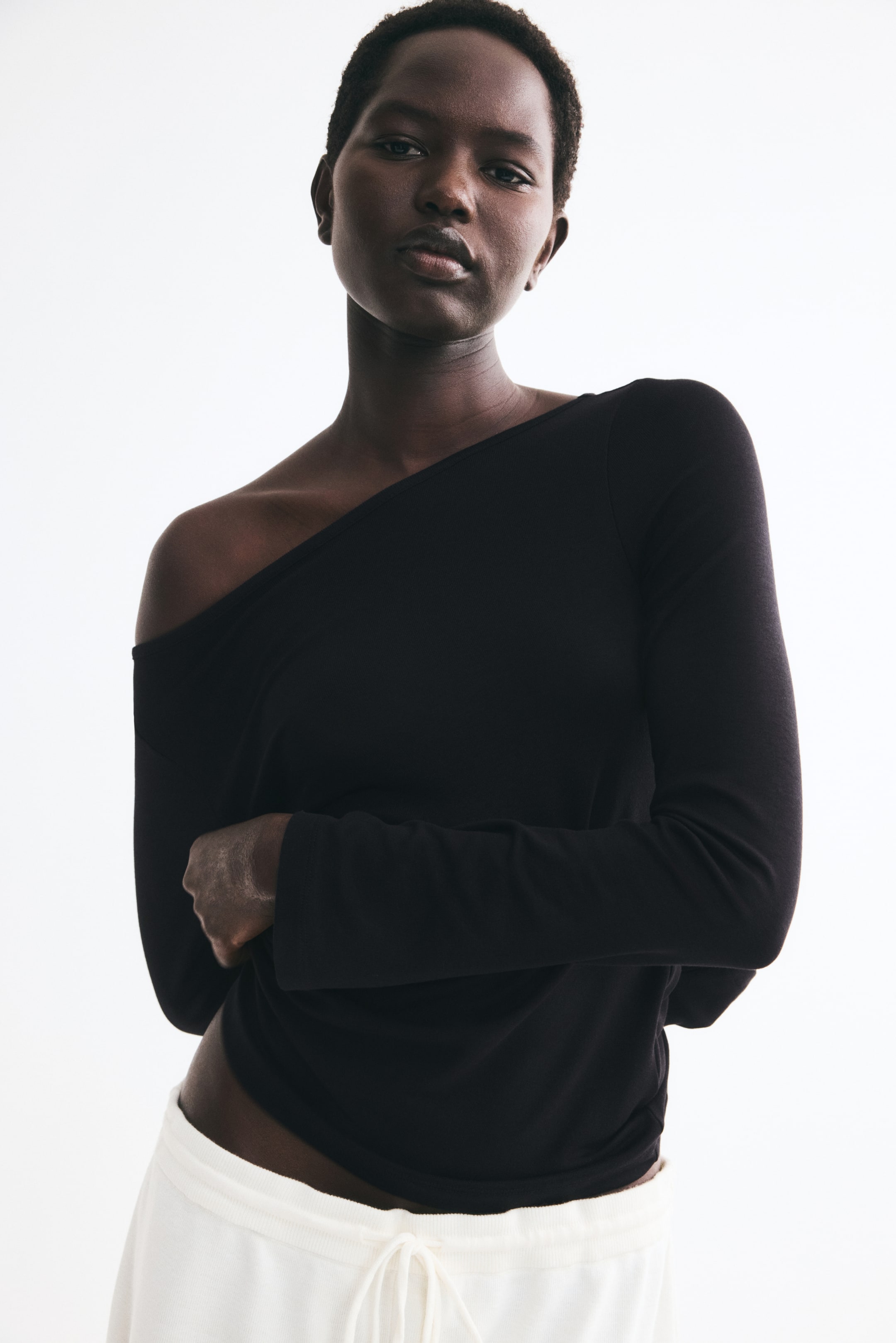 View larger image: The model is wearing a black long-sleeved fine-knit top, styled with the boat neckline pulled down off one shoulder. The top is paired with cream-colored drawstring trousers, visible at the waist.