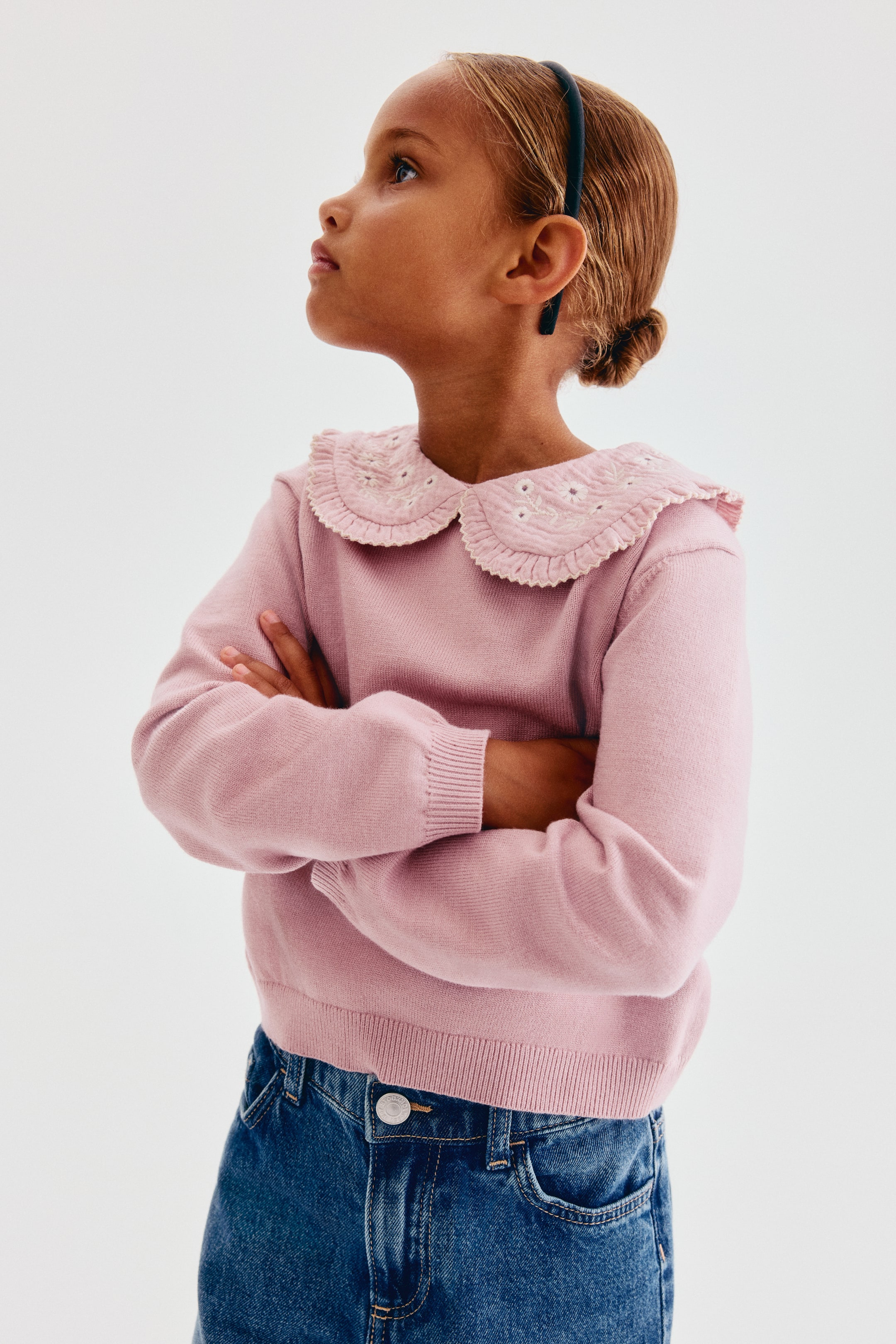 View larger image: A pink jumper in a soft, fine knit is shown from the front, featuring a white embroidered Peter Pan collar with frill trim. The jumper is designed with gently dropped shoulders, long sleeves with ribbed cuffs, and a ribbed hem, and is styled with blue denim jeans.