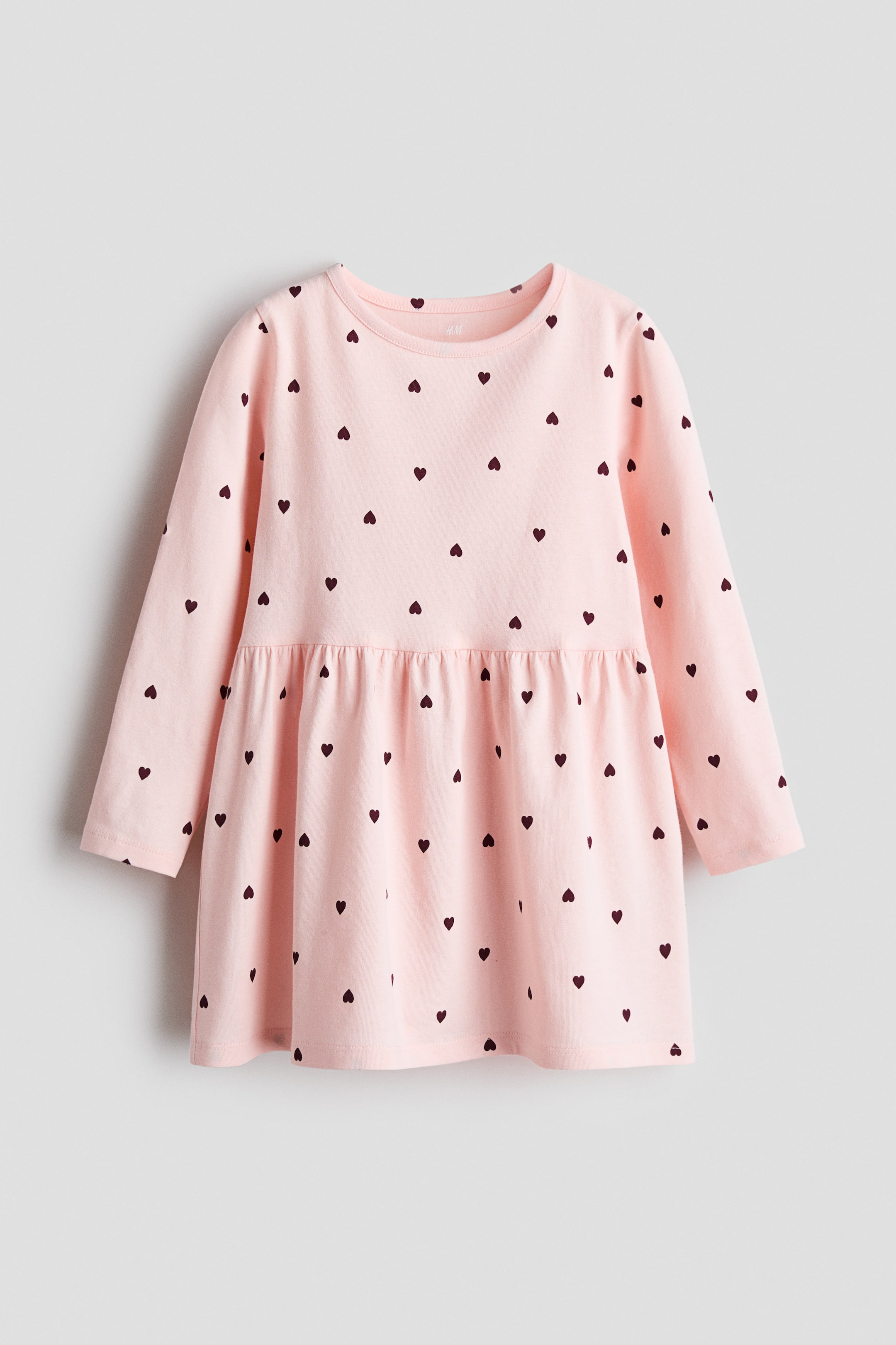 View larger image: A pink children's dress is showcasing its front view, featuring long sleeves, a round neckline, and an all-over pattern of small dark hearts, with a gathered seam creating a flared skirt.