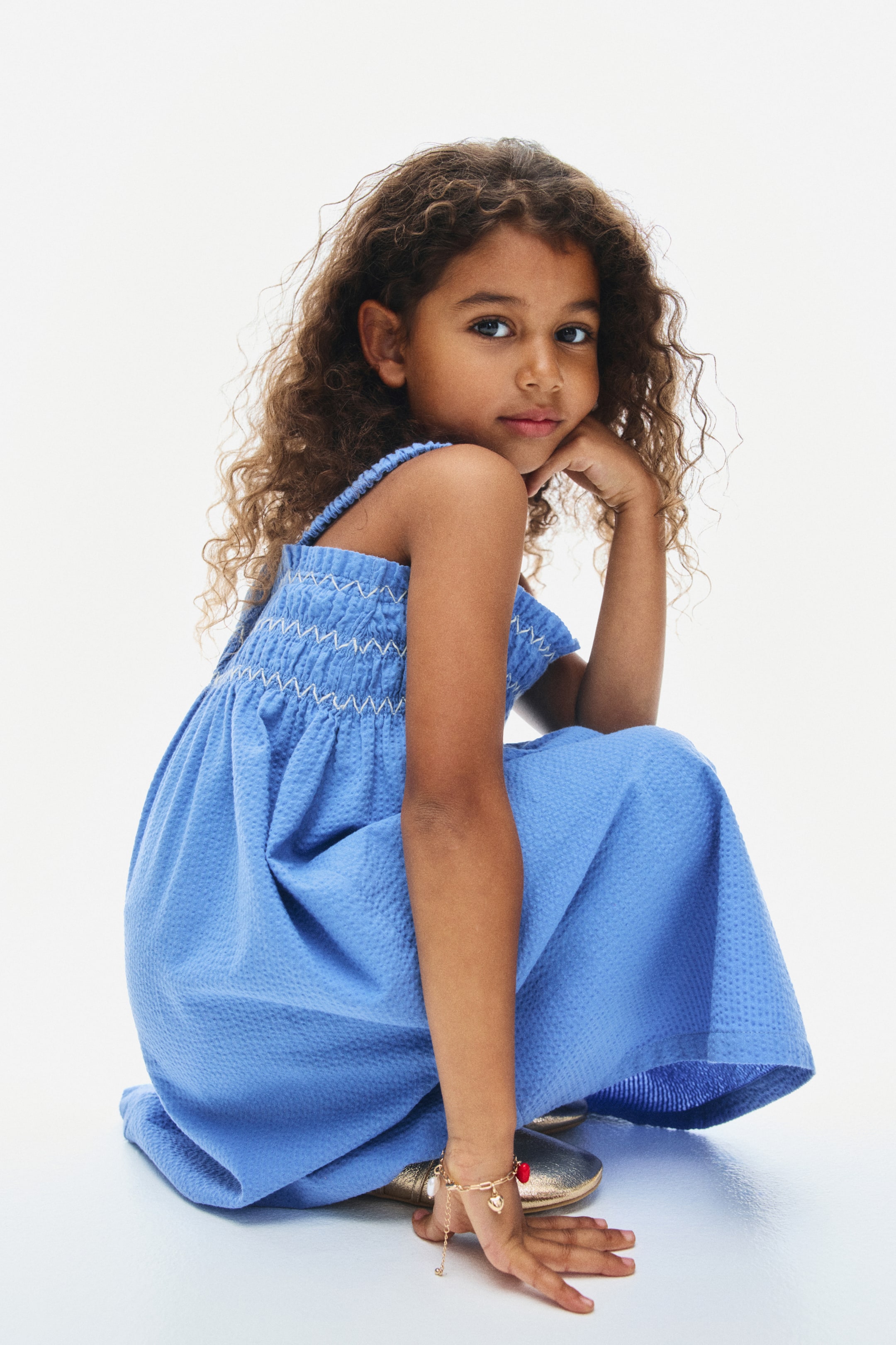 View larger image: A blue seersucker dress is shown with a smocked bodice featuring white zigzag stitching and elasticated shoulder straps. The dress flows into a flared skirt, showcasing its textured fabric as the model crouches and faces left.