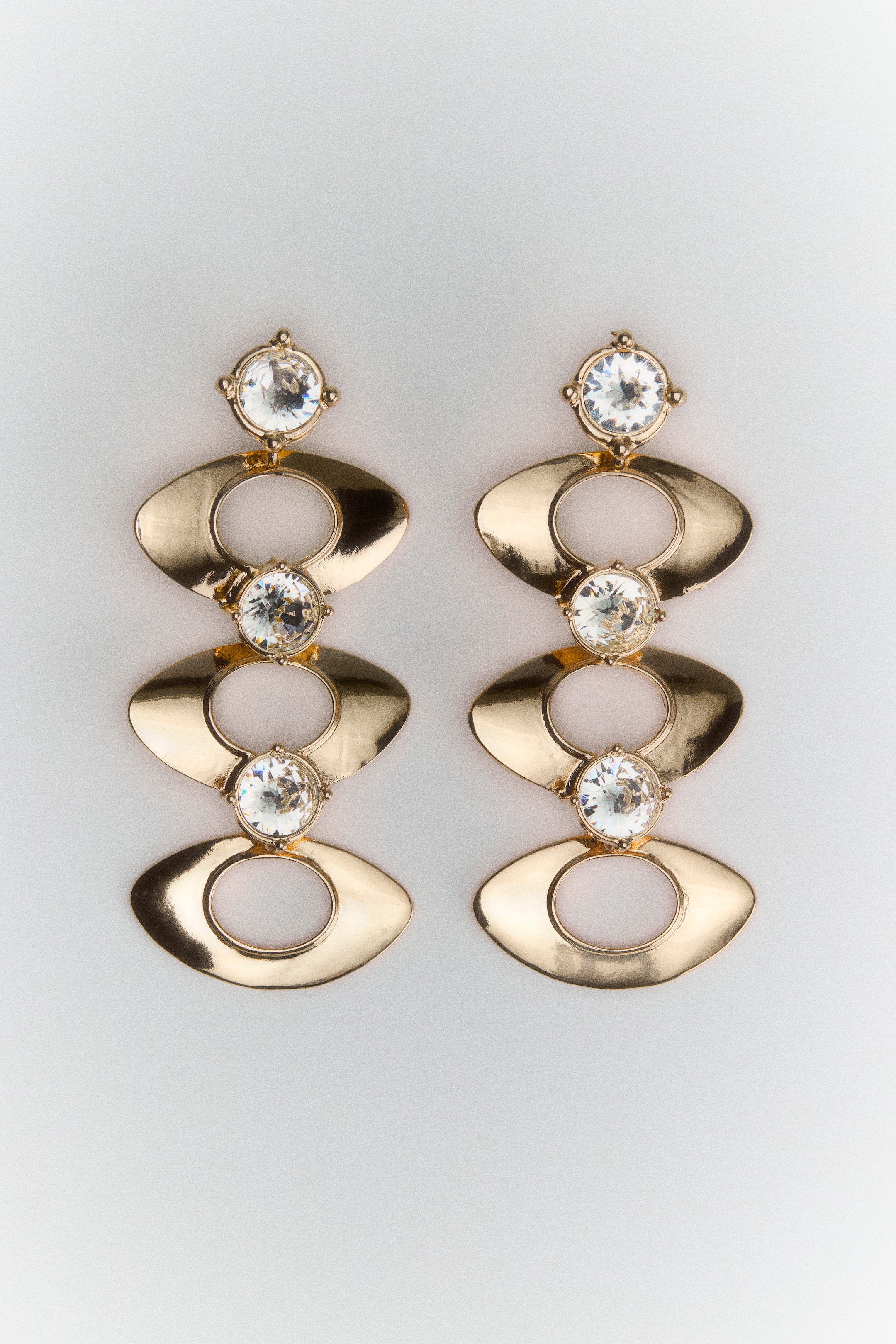 View larger image: A pair of gold-toned metal dangle earrings is shown from the front, featuring a vertical drop design composed of three round, clear glass rhinestones alternating with three oval-shaped metal pendants with large central cut-outs.