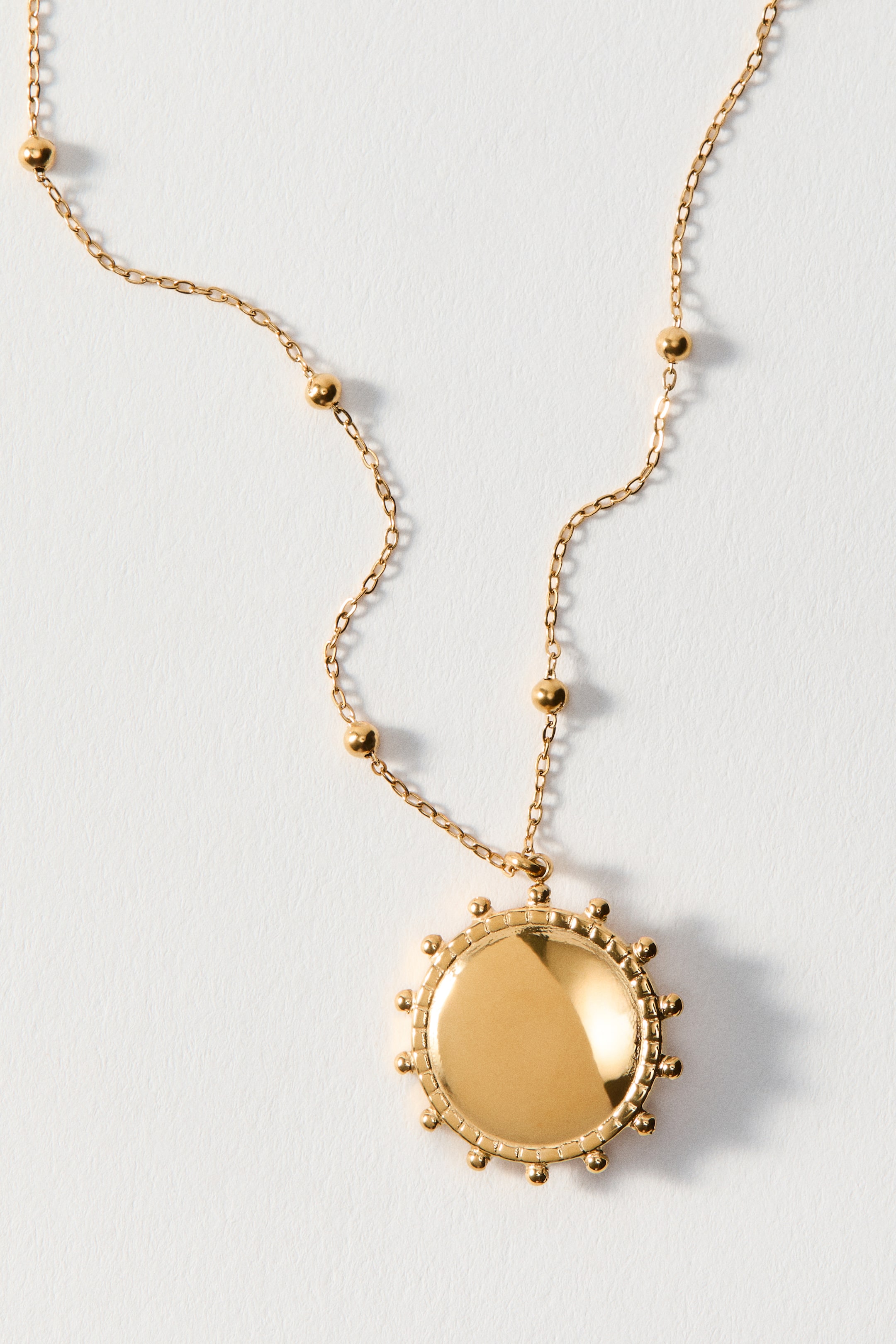 Agrandir l'image: Un collier doré présente une chaîne à maillons qui mène à un pendentif en forme d'éclat de soleil. Le pendentif affiche un centre lisse et réfléchissant, entouré de petites protubérances sphériques le long de son bord.