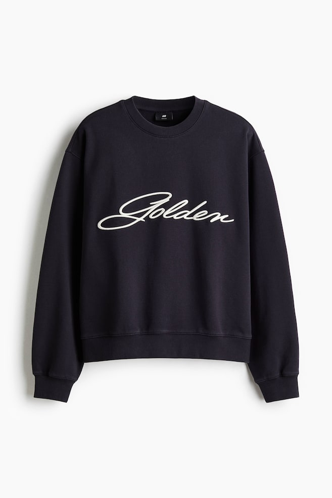 濃い青のスウェットシャツは、胸に白い刺繍の「Golden」スクリプトが施されたFRONTを見せています。丸いリブ付きのネックライン、ドロップショルダー、長袖、幅広のリブ付きカフスと裾がデザインされており、ゆったりとしたフィット感を演出しています。 - 1
