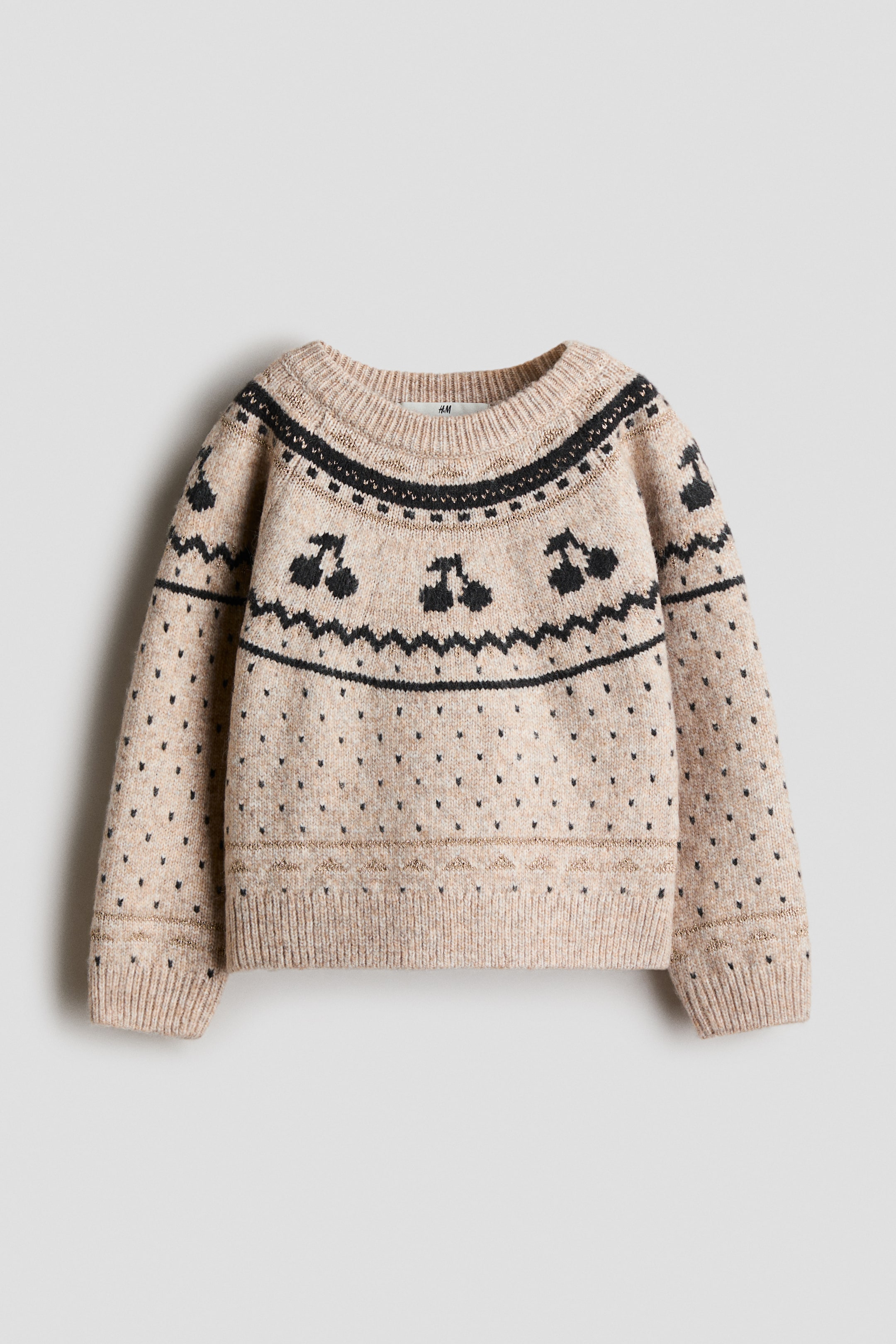 View larger image: A beige jacquard-knit sweater is shown from the front, featuring a round ribbed neckline, long raglan sleeves, and ribbed cuffs and hem. The sweater displays a black knit pattern of cherries and geometric shapes across the yoke, with small black specks throughout the body and subtle metallic stripes accenting the lower hem and cuffs.