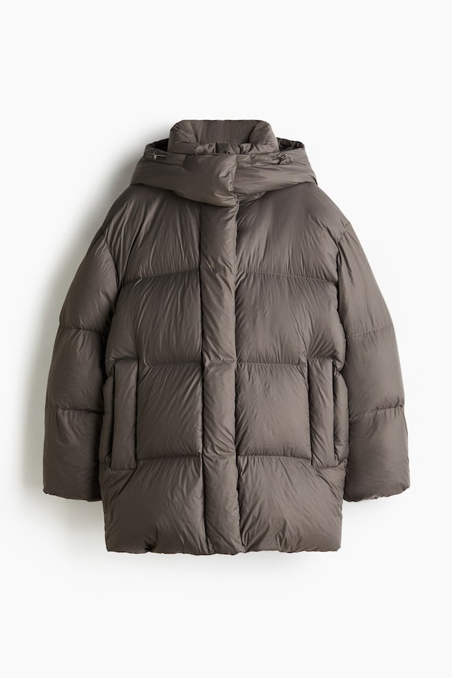An oversized puffer jacket in dark taupe nylon with down and feather filling is presented from the FRONT. It is featuring a detachable hood with an elasticated drawstring and cord stoppers, a stand collar, and a front wind flap concealing a zip and press-studs. The jacket is also displaying welt side pockets with concealed zips and an elasticated drawstring with cord stoppers at the hem. - 1