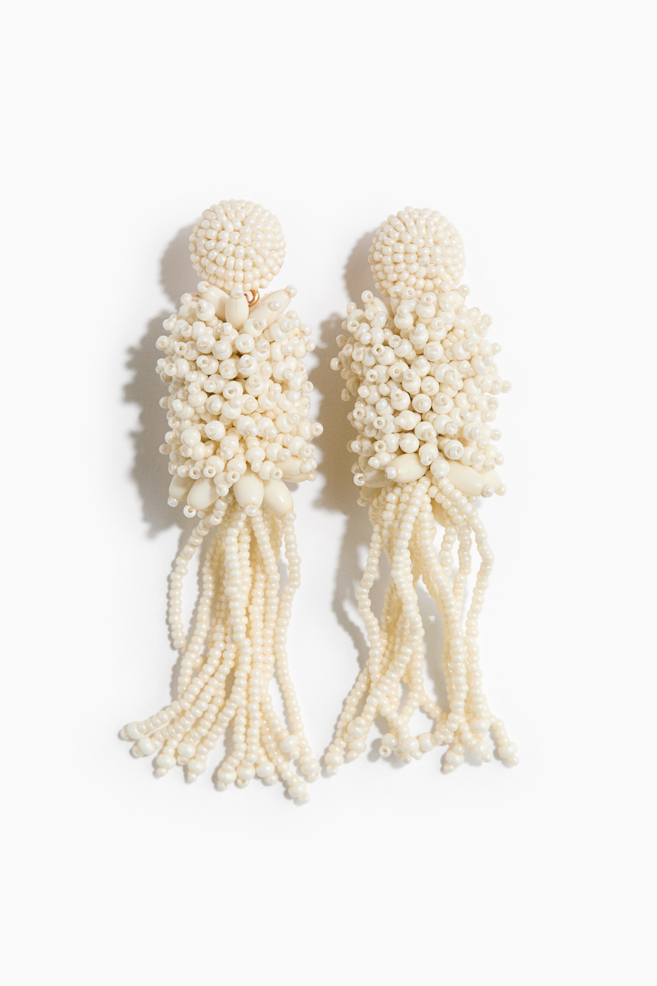 View larger image: A pair of cream-coloured beaded earrings featuring a dome-shaped top section, densely adorned with small beads, connected by a gold-toned hook to a cylindrical main body. The main body is similarly adorned with small beads, interspersed with several larger, elongated beads. Multiple strands of small beads hang from its base, creating a flowing, tassel-like fringe.