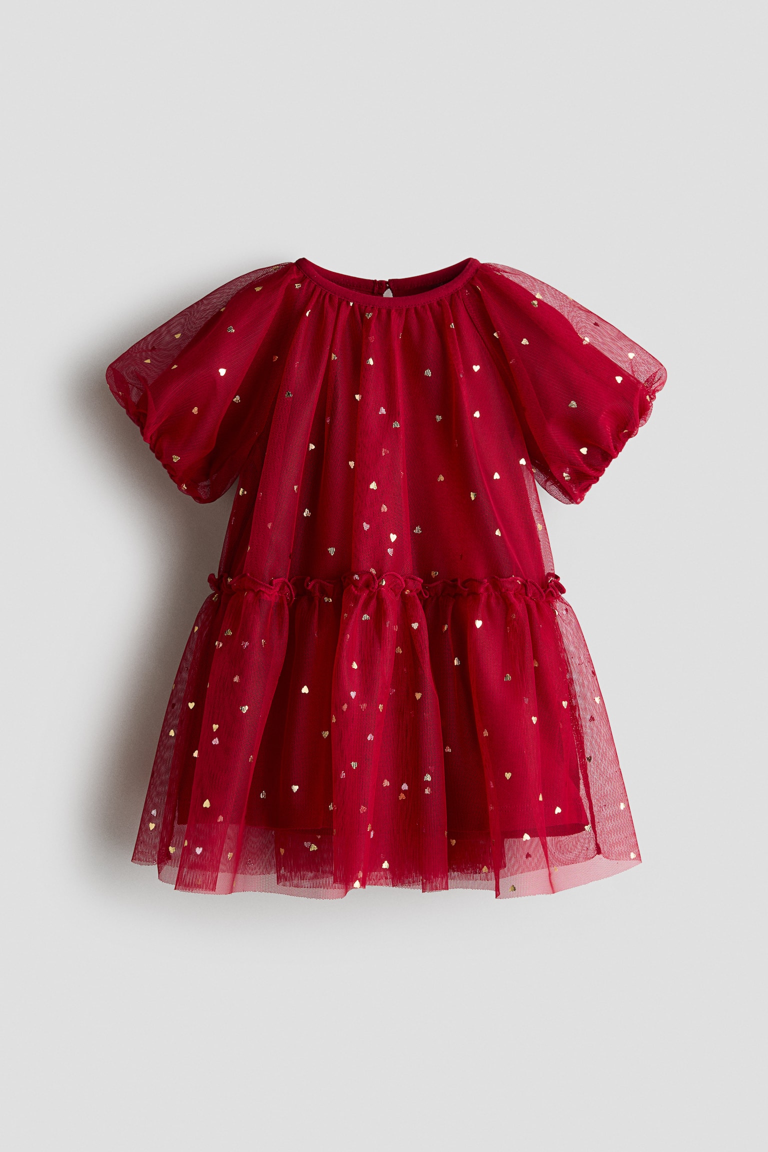 A red childrenâs dress is shown from the front, featuring a sheer tulle overlay patterned with small metallic gold hearts. The dress is designed with a round neckline, short puff sleeves, and a gathered, tiered skirt with a ruffled waist seam.