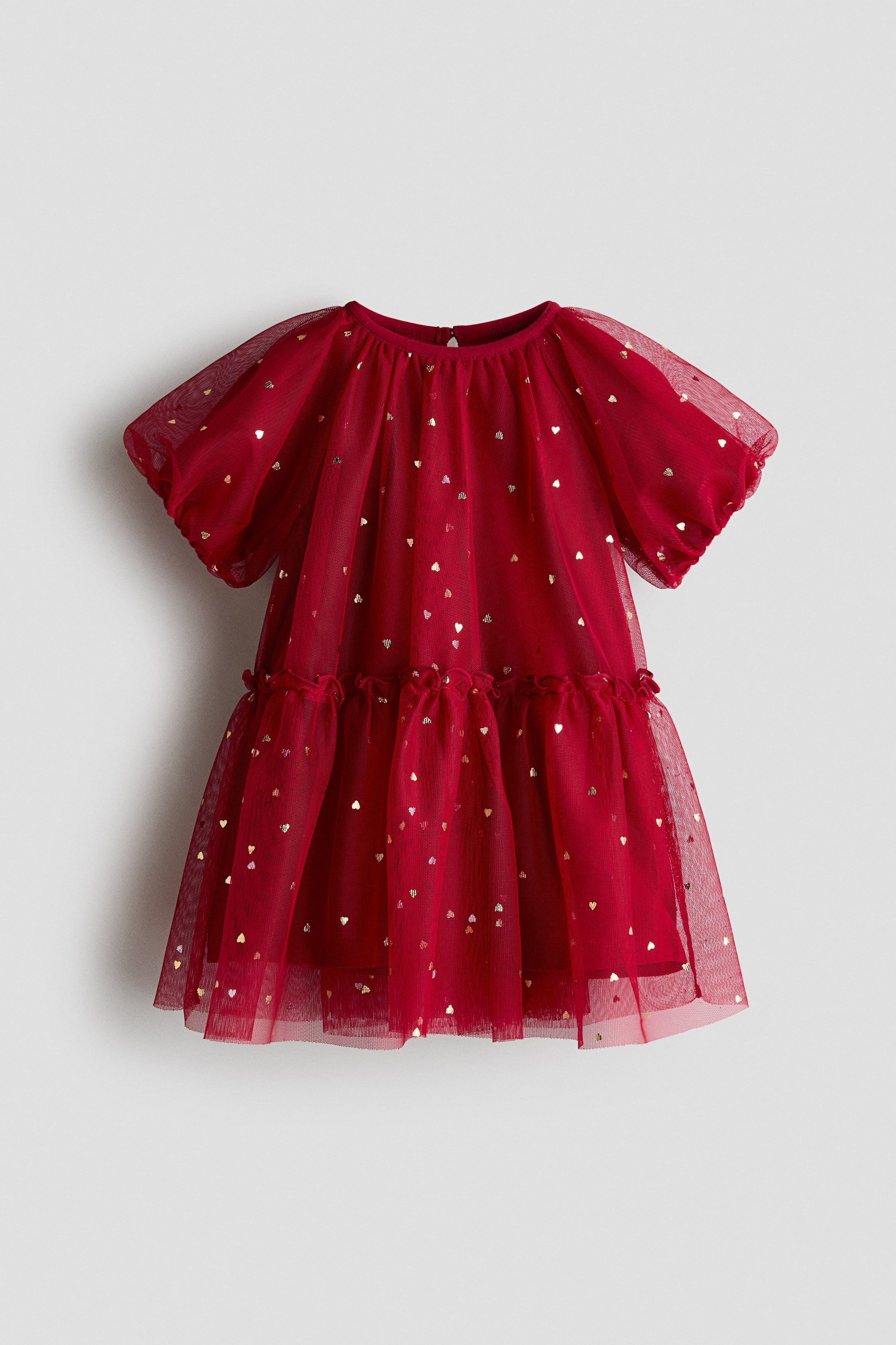 View larger image: A red children’s dress is shown from the front, featuring a sheer tulle overlay patterned with small metallic gold hearts. The dress is designed with a round neckline, short puff sleeves, and a gathered, tiered skirt with a ruffled waist seam.
