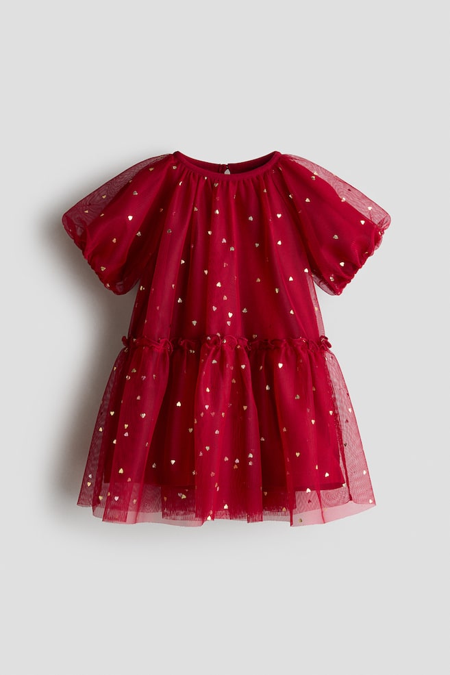 A red childrenâs dress is shown from the front, featuring a sheer tulle overlay patterned with small metallic gold hearts. The dress is designed with a round neckline, short puff sleeves, and a gathered, tiered skirt with a ruffled waist seam. - 1